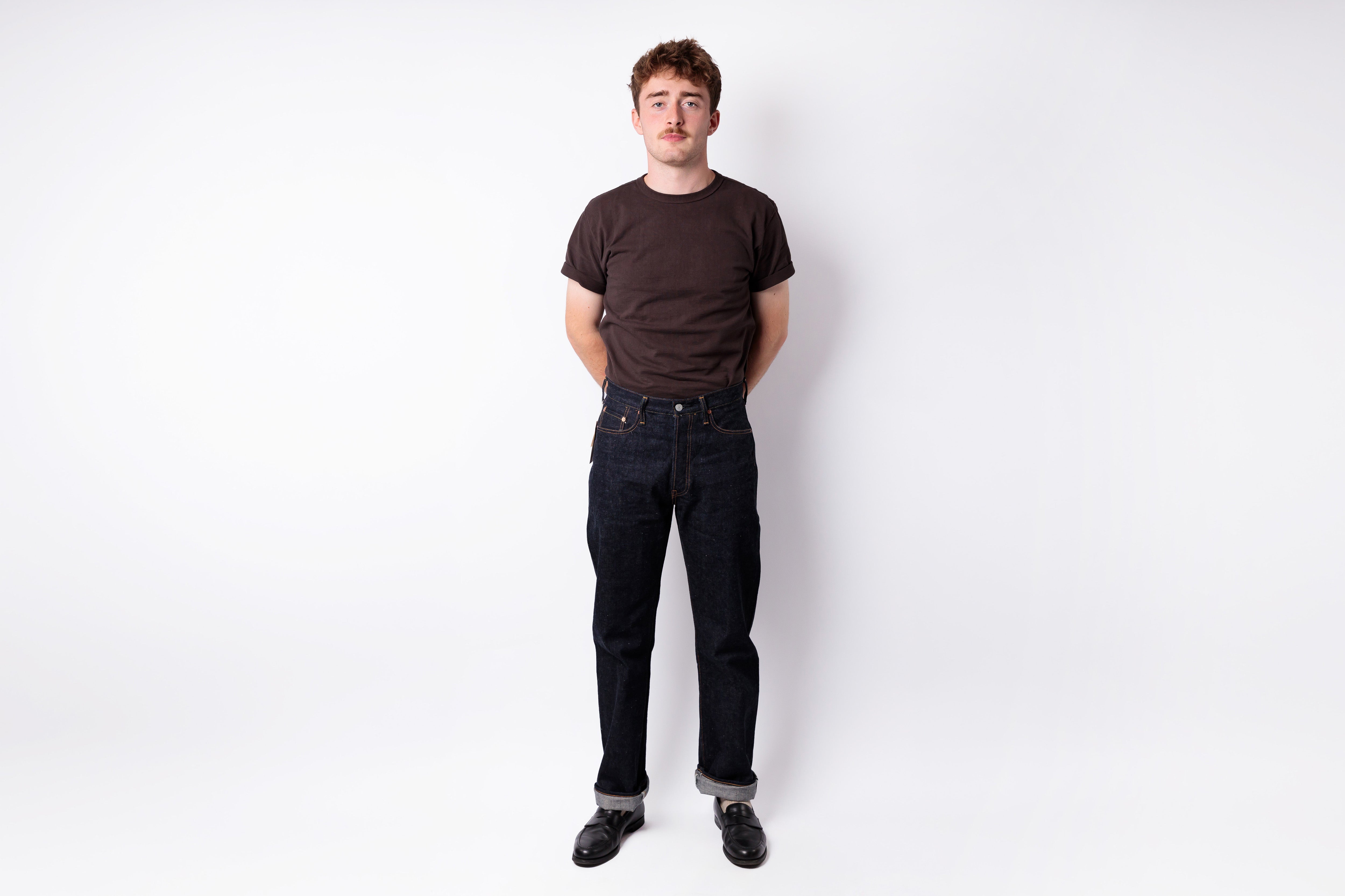 A young man standing against a white studio background, wearing a dark brown crewneck t-shirt, dark blue jeans, and black shoes.