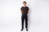 A young man standing against a white studio background, wearing a dark brown crewneck t-shirt, dark blue jeans, and black shoes.