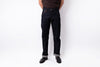 A person wearing dark indigo denim jeans with rolled cuffs, paired with a brown shirt and black shoes, standing against a plain white studio background.