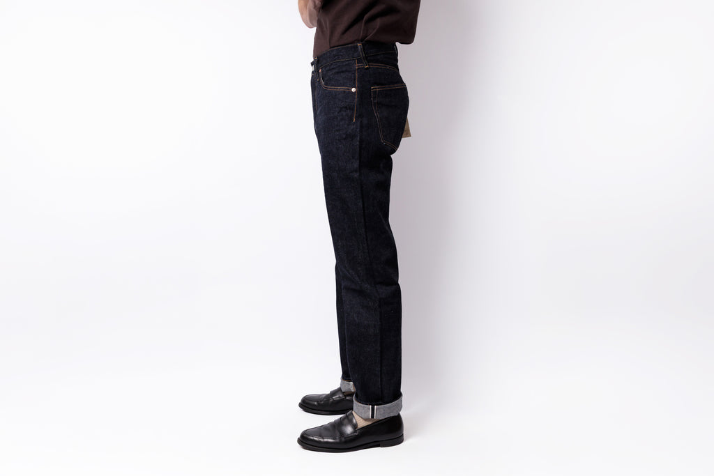 A person shown from the waist down wearing dark indigo slim-fit jeans with rolled cuffs, black leather shoes, and a dark brown top, standing against a plain white studio background.