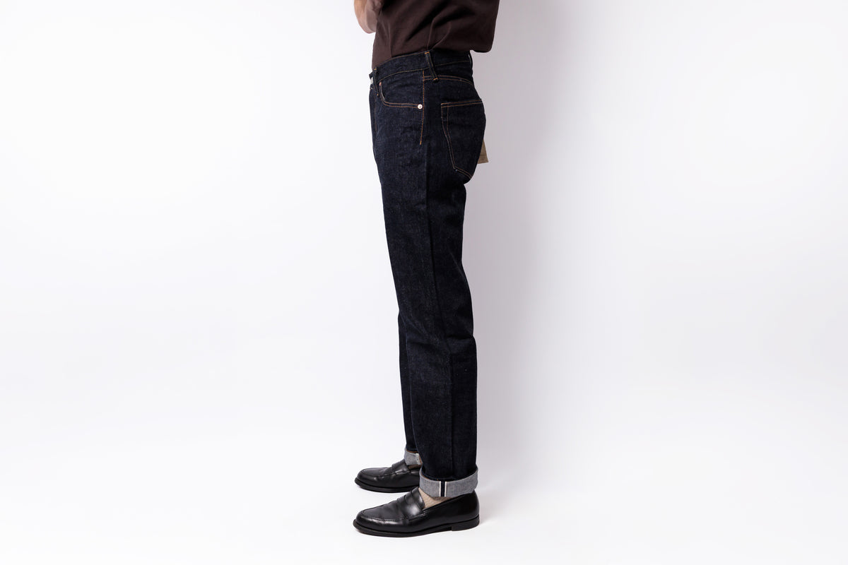 A person shown from the waist down wearing dark indigo slim-fit jeans with rolled cuffs, black leather shoes, and a dark brown top, standing against a plain white studio background.