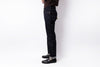 A person shown from the waist down wearing dark indigo slim-fit jeans with rolled cuffs, black leather shoes, and a dark brown top, standing against a plain white studio background.