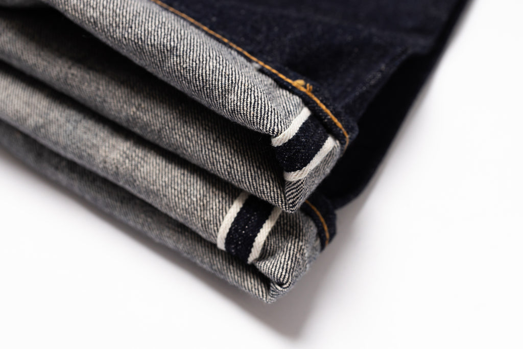A close-up of folded dark indigo denim jeans showing the twill fabric texture, contrast orange stitching, and white and black stripe detail along the hem, set against a clean white background.