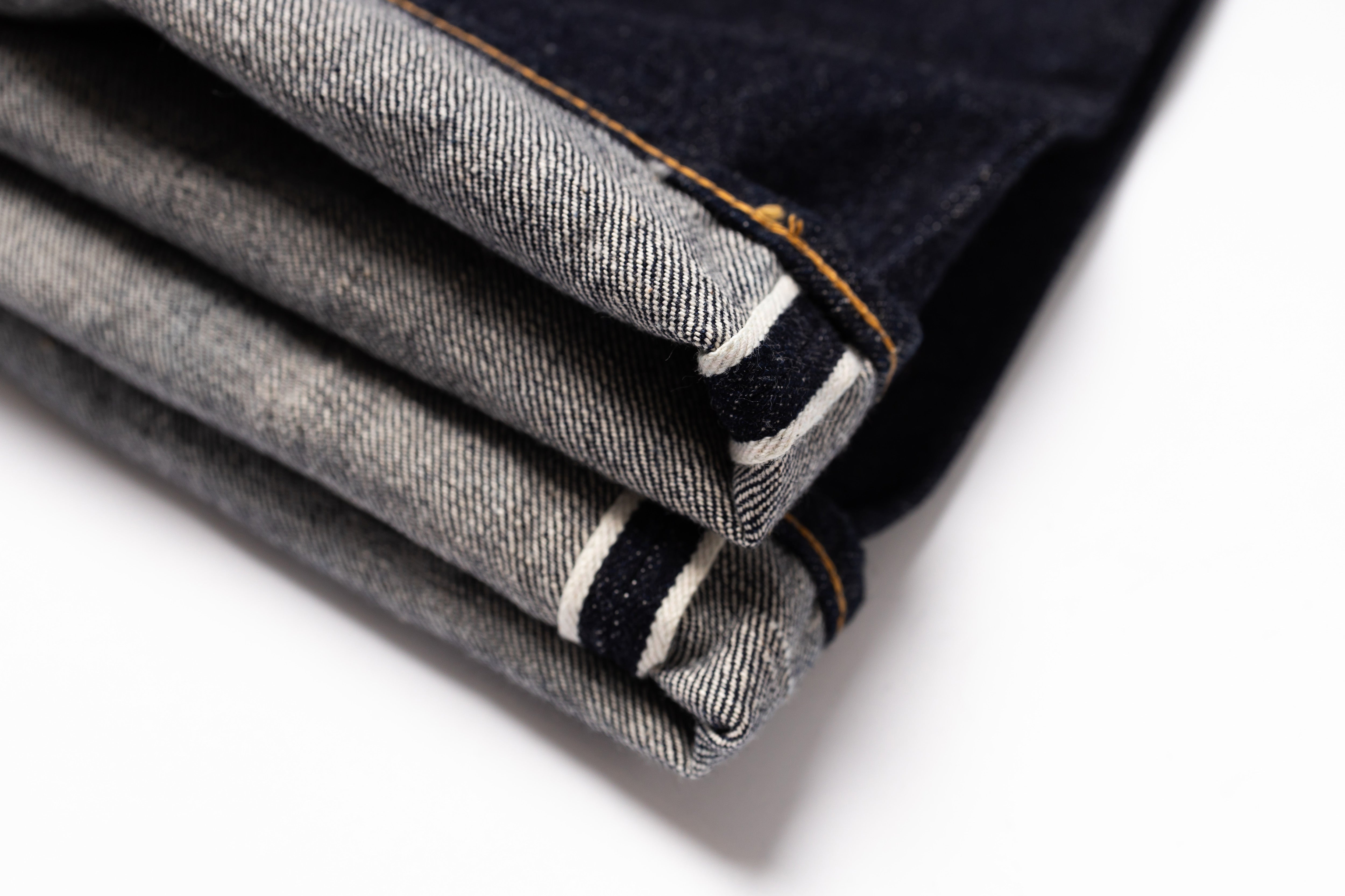 A close-up of folded dark indigo denim jeans showing the twill fabric texture, contrast orange stitching, and white and black stripe detail along the hem, set against a clean white background.