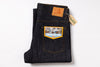 A folded pair of dark indigo denim jeans with a distinctive yellow and blue patch on the back pocket, featuring a brown leather patch and a paper hangtag on a white background.