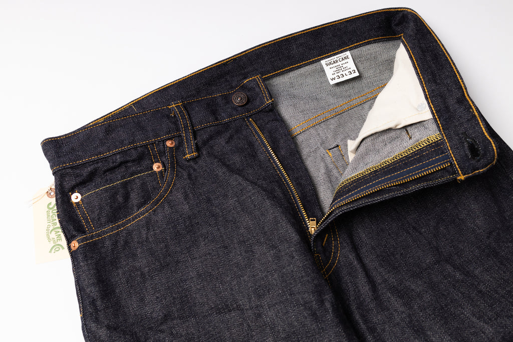 Close-up of dark indigo denim jeans laid flat on a white background, showing the waistband, front pocket, button, zipper, and interior label.