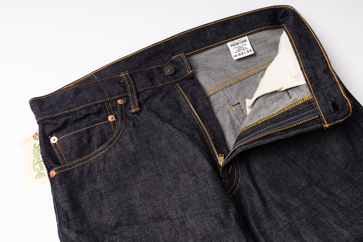Close-up of dark indigo denim jeans laid flat on a white background, showing the waistband, front pocket, button, zipper, and interior label.