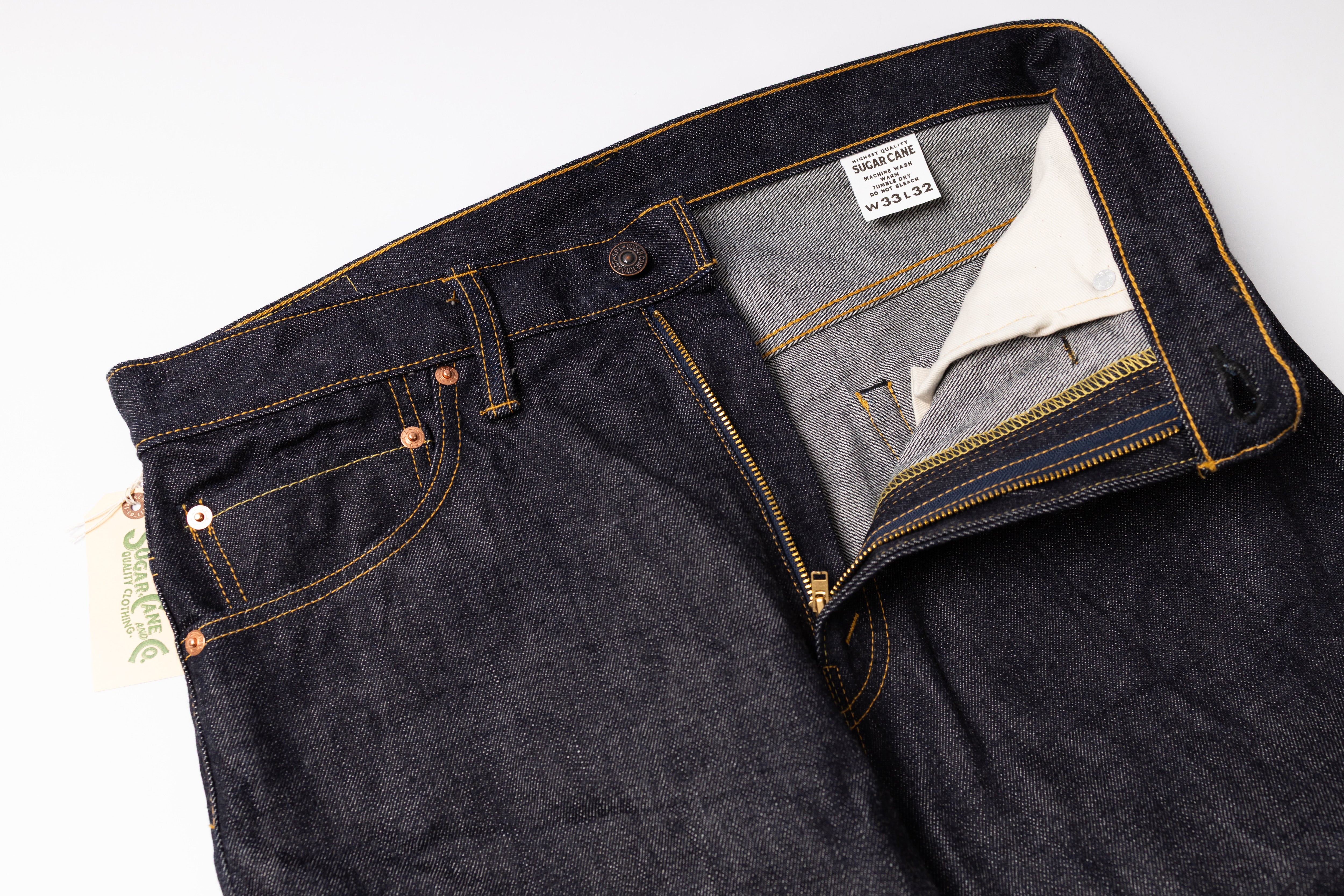 Close-up of dark indigo denim jeans laid flat on a white background, showing the waistband, front pocket, button, zipper, and interior label.