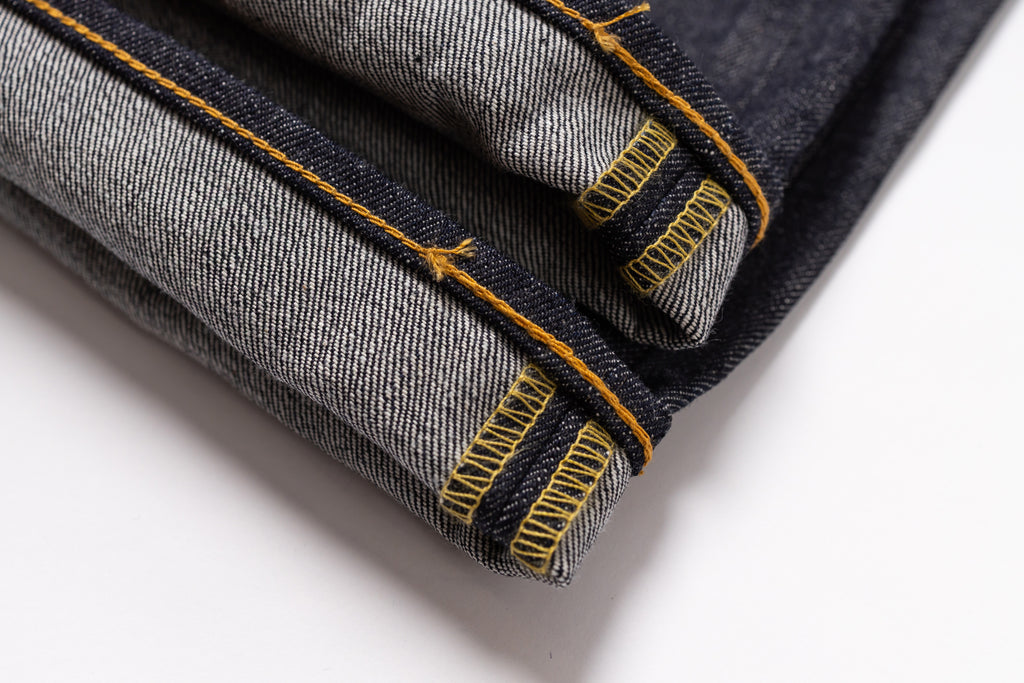 Close-up of folded dark indigo denim jeans showing the twill fabric texture and bright yellow contrast stitching on a white background.