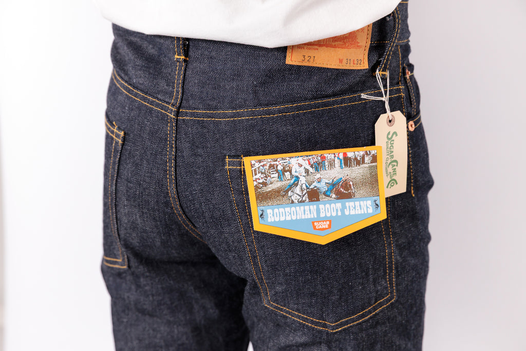 Close-up of the back waistband and pocket area of dark indigo denim jeans, showing a tan leather patch, a paper hangtag, and a decorative back pocket label reading 'Buckman Boot Jeans'.