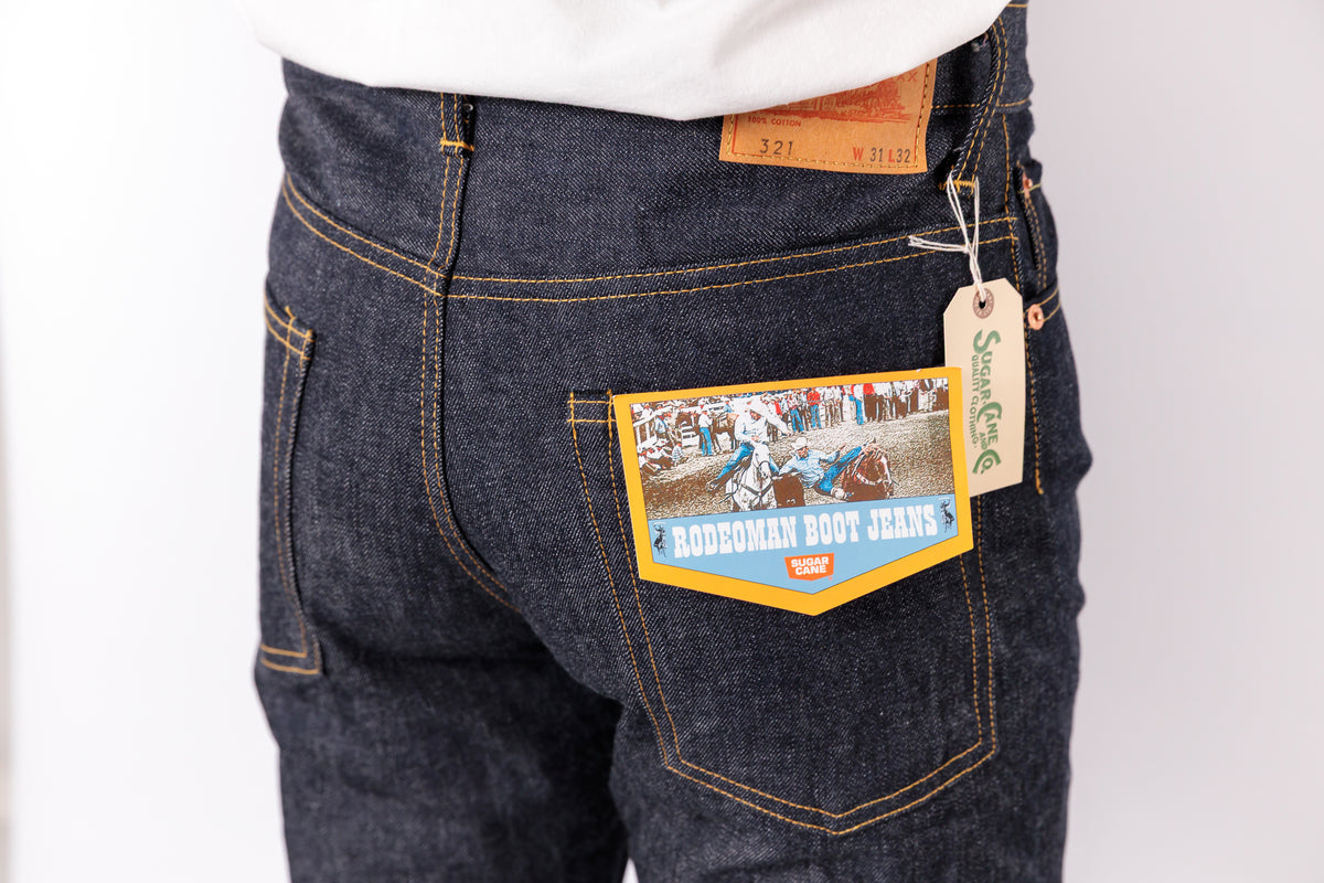 Close-up of the back waistband and pocket area of dark indigo denim jeans, showing a tan leather patch, a paper hangtag, and a decorative back pocket label reading 'Buckman Boot Jeans'.