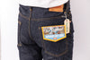 Close-up of the back waistband and pocket area of dark indigo denim jeans, showing a tan leather patch, a paper hangtag, and a decorative back pocket label reading 'Buckman Boot Jeans'.