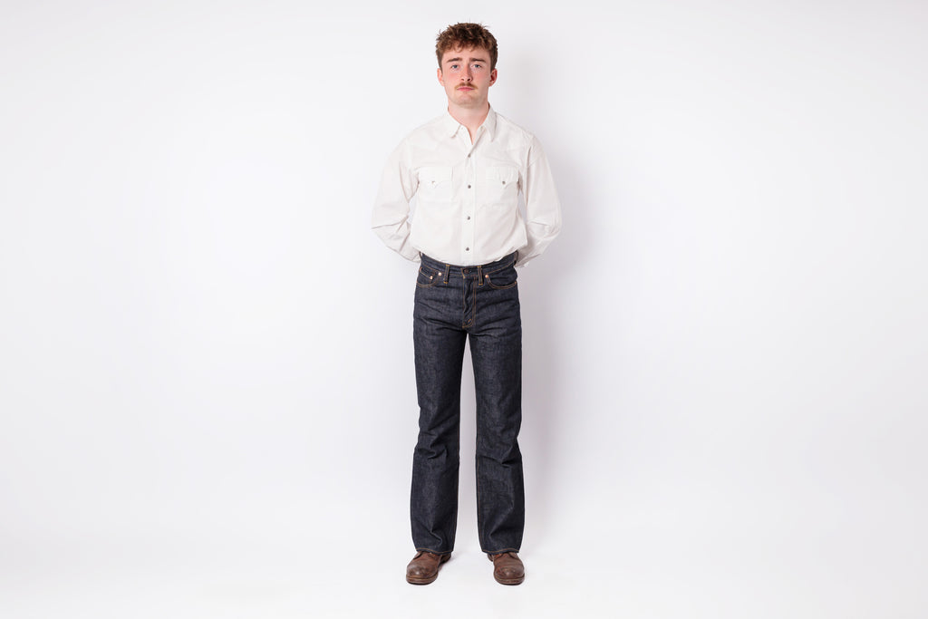A man standing against a plain white studio background, wearing a light cream long-sleeve button-up shirt, dark blue straight-leg jeans, and brown shoes.