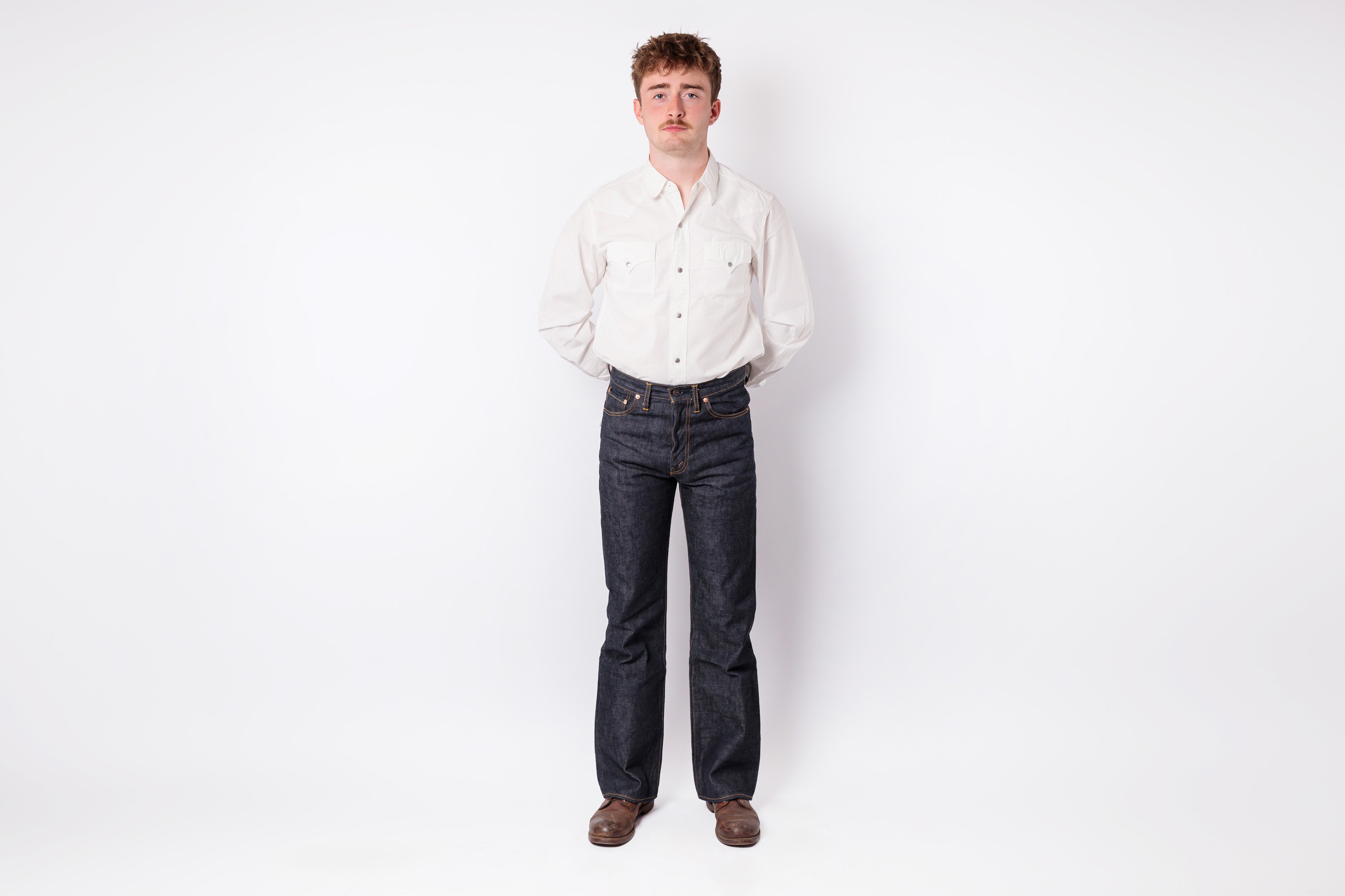 A man standing against a plain white studio background, wearing a light cream long-sleeve button-up shirt, dark blue straight-leg jeans, and brown shoes.