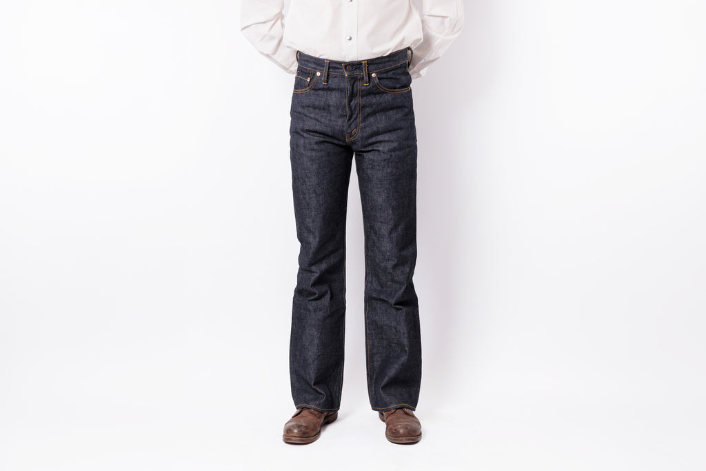 A person wearing dark indigo denim jeans, a white button-down shirt, and brown leather shoes, standing against a plain white studio background.