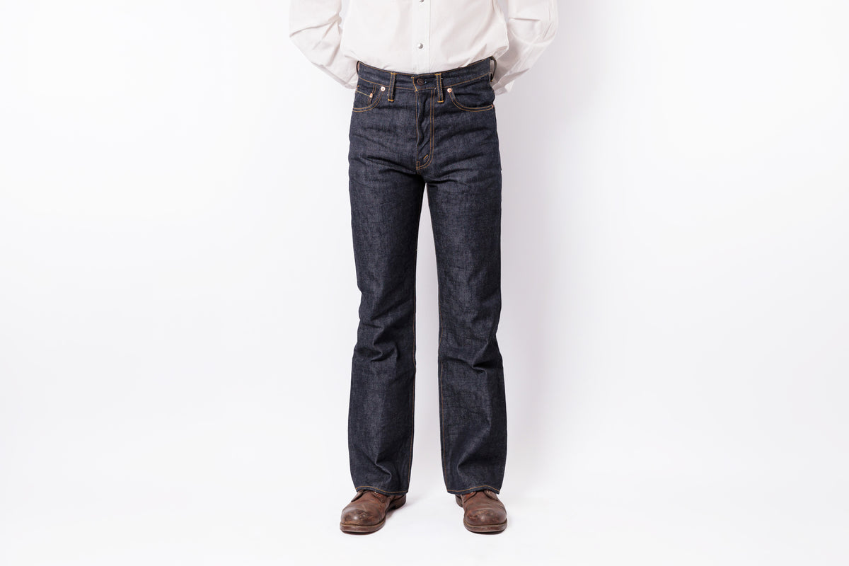 A person wearing dark indigo denim jeans, a white button-down shirt, and brown leather shoes, standing against a plain white studio background.
