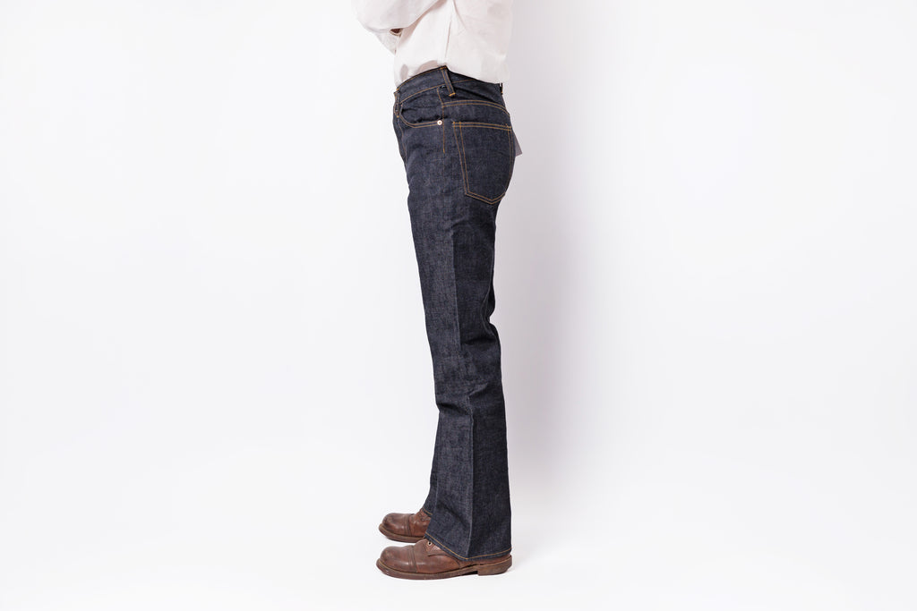 A person shown from the waist down wearing dark indigo flare jeans, a white shirt, and brown leather shoes, standing against a plain white studio background.