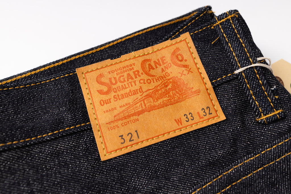 Close-up of dark indigo denim jeans showing the interior waistband and a prominent tan leather patch with the Sugar Creek 6 logo and text.