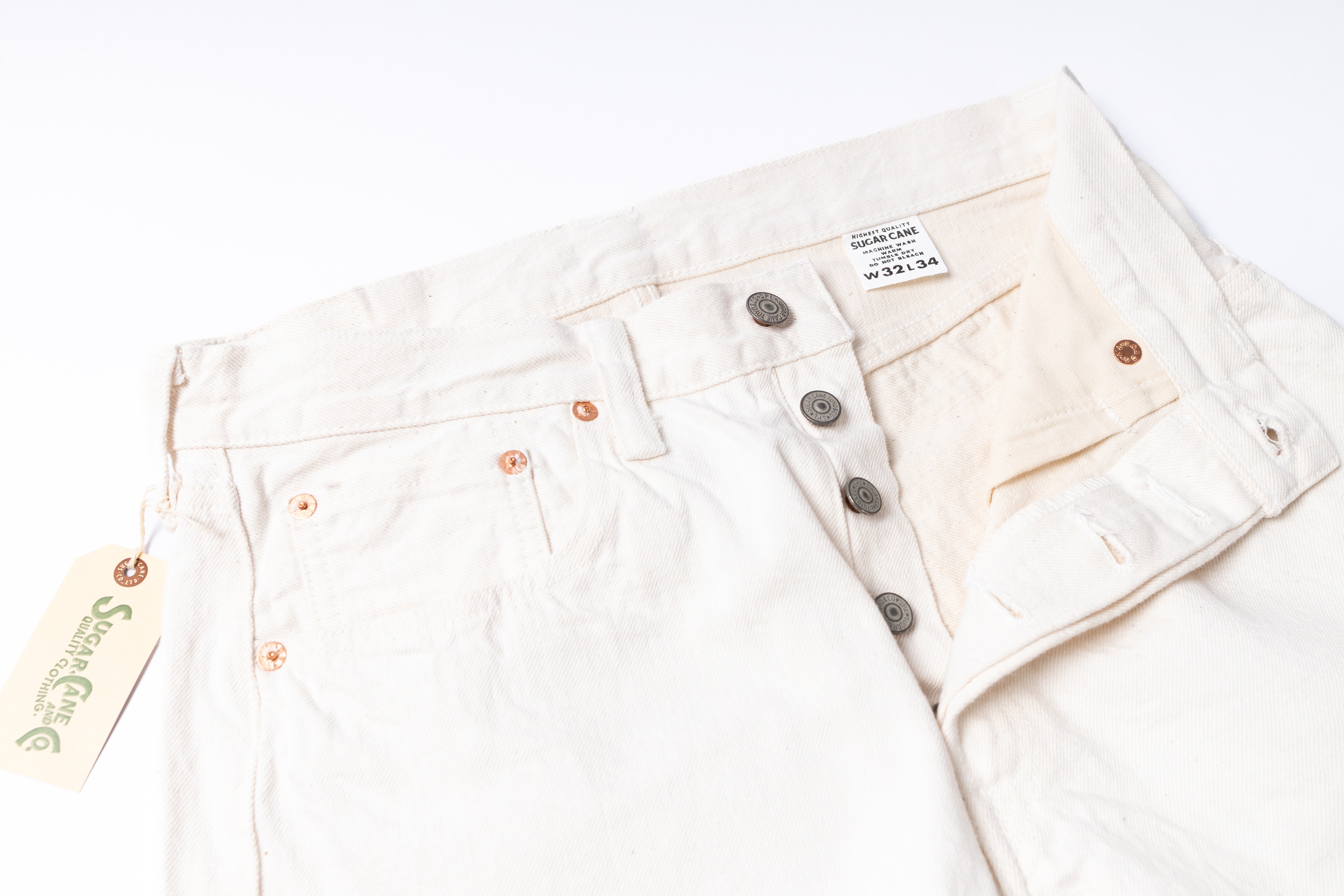 A close-up of off-white denim jeans laid flat on a white background, showing the waistband, button fly, front pockets, and a hanging tag with  branding.