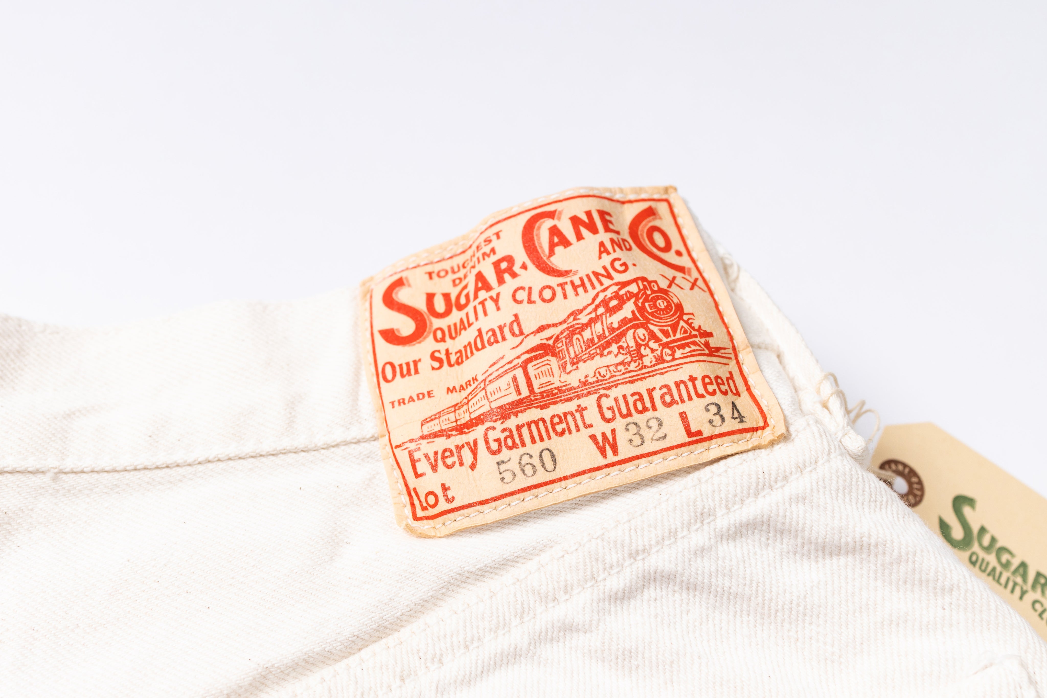 A close-up of a vintage cream-colored cotton garment label sewn onto white fabric, featuring red printed text that reads 'Sugar-Cane An Quality Clothing Co.' with a train illustration.