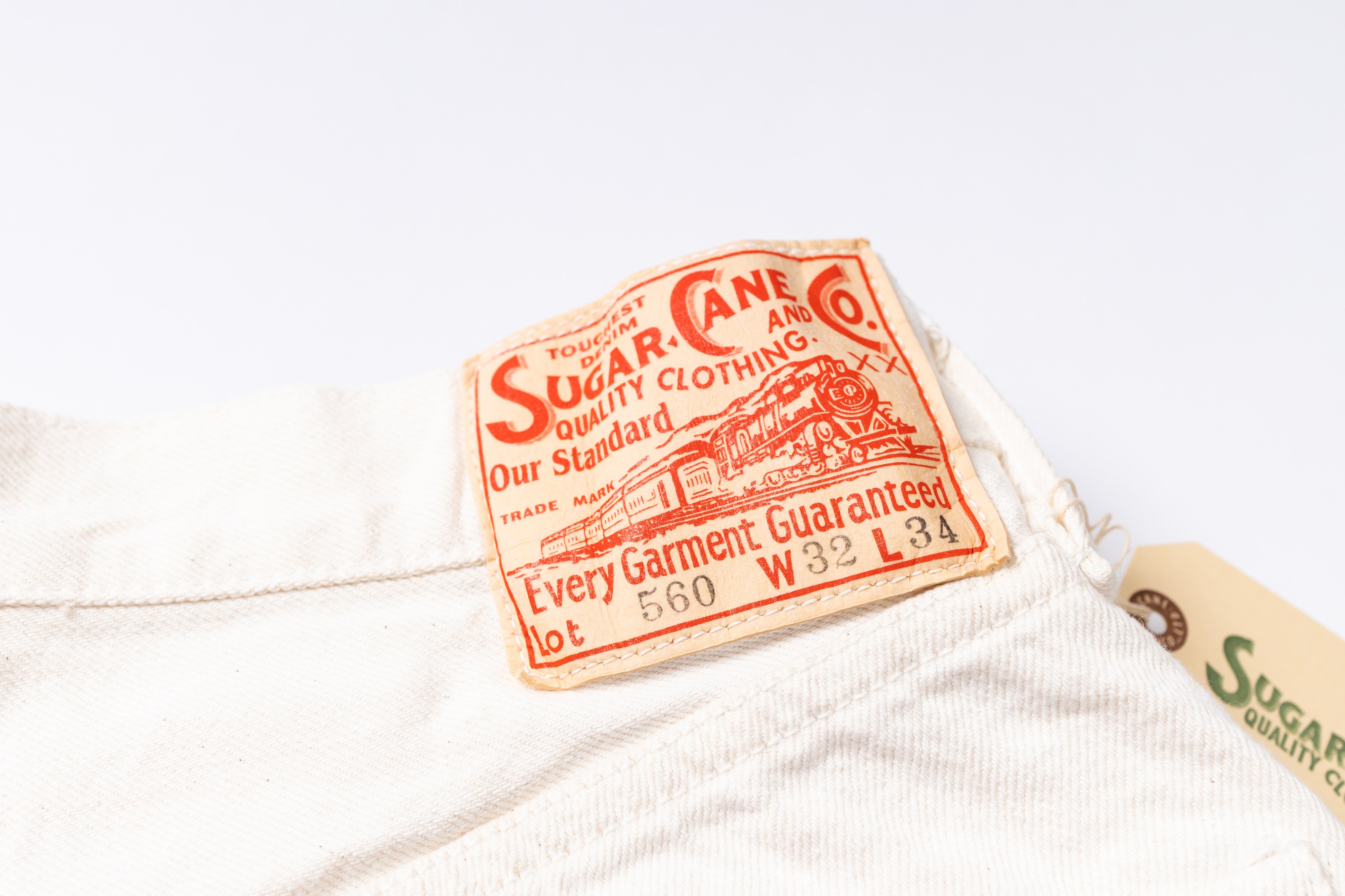 A close-up of a vintage cream-colored cotton garment label sewn onto white fabric, featuring red printed text that reads 'Sugar-Cane An Quality Clothing Co.' with a train illustration.