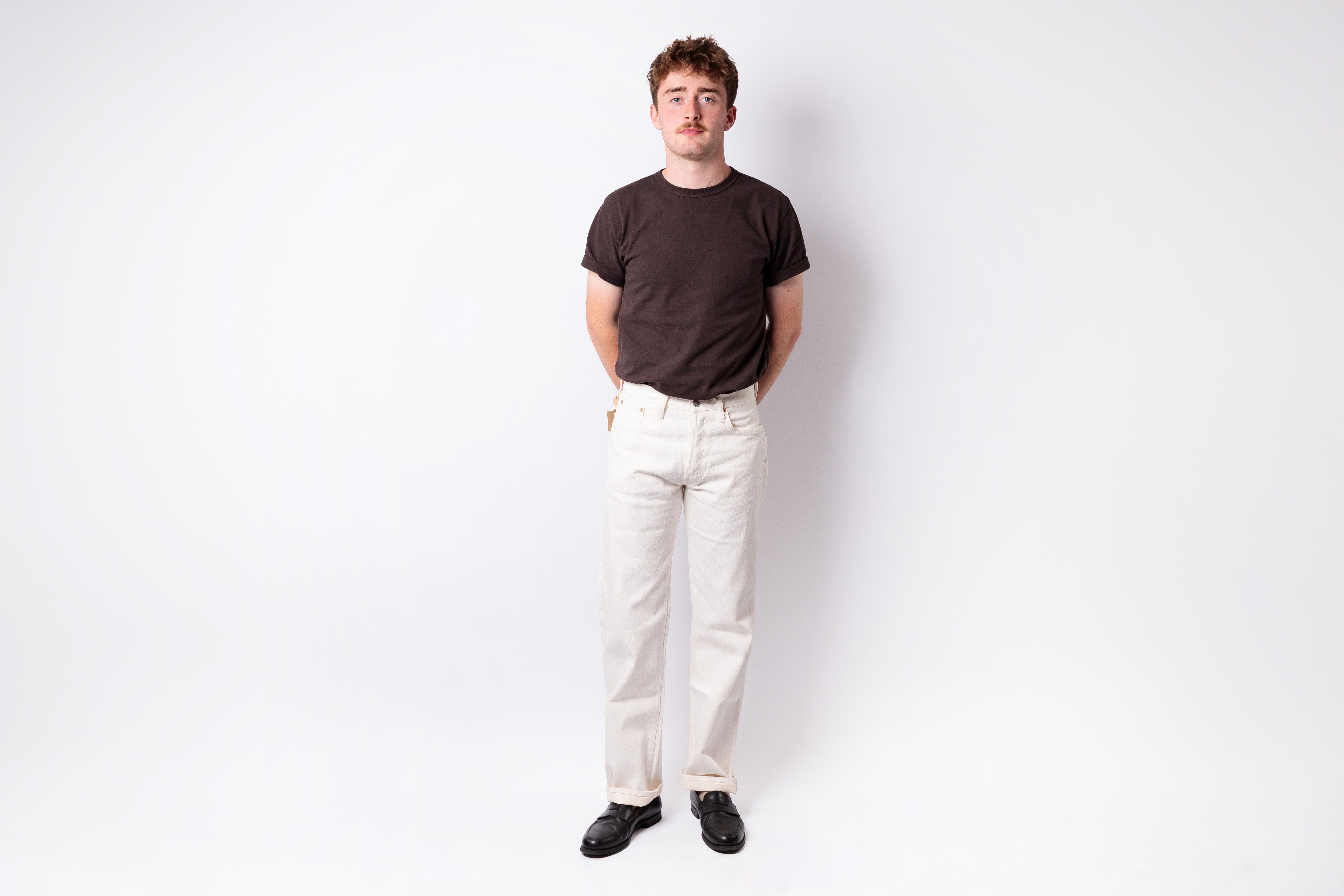 A man standing against a plain white studio background, wearing a dark brown short-sleeve crewneck t-shirt, off-white trousers, and black shoes.