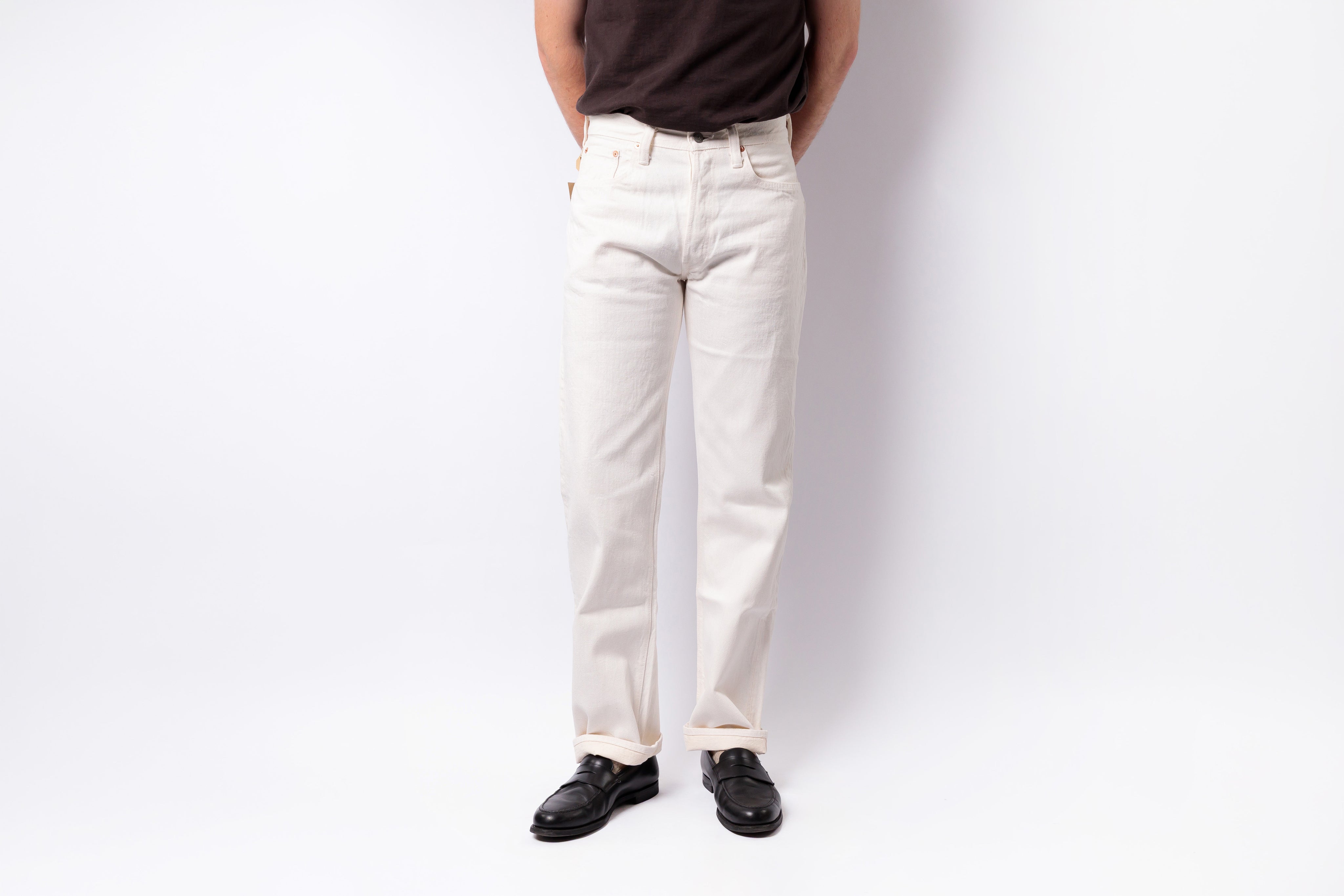 A person wearing off-white straight-leg jeans, a dark brown t-shirt, and black leather shoes, standing against a plain white studio background.