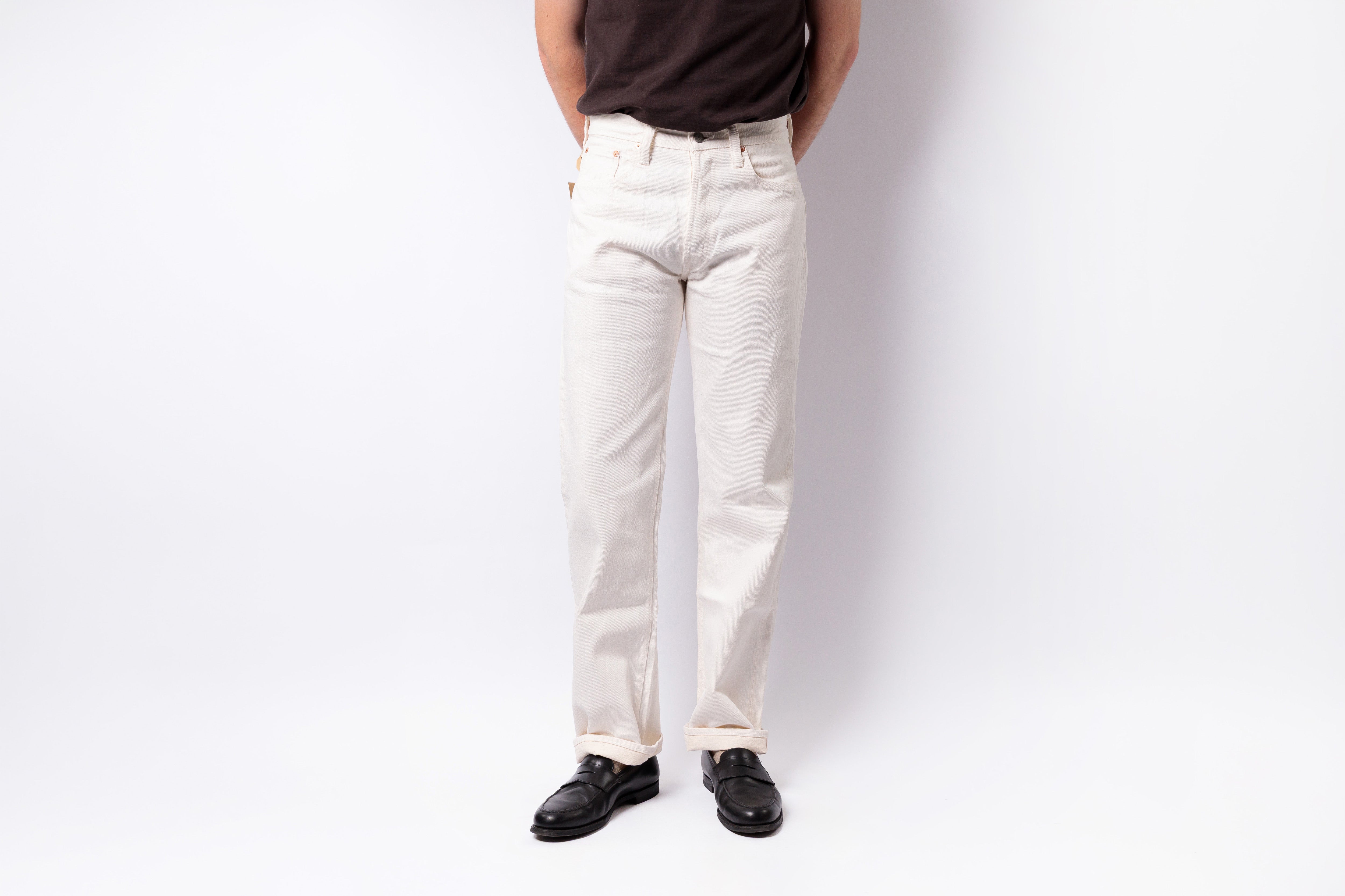 A person wearing off-white straight-leg jeans, a dark brown t-shirt, and black leather shoes, standing against a plain white studio background.