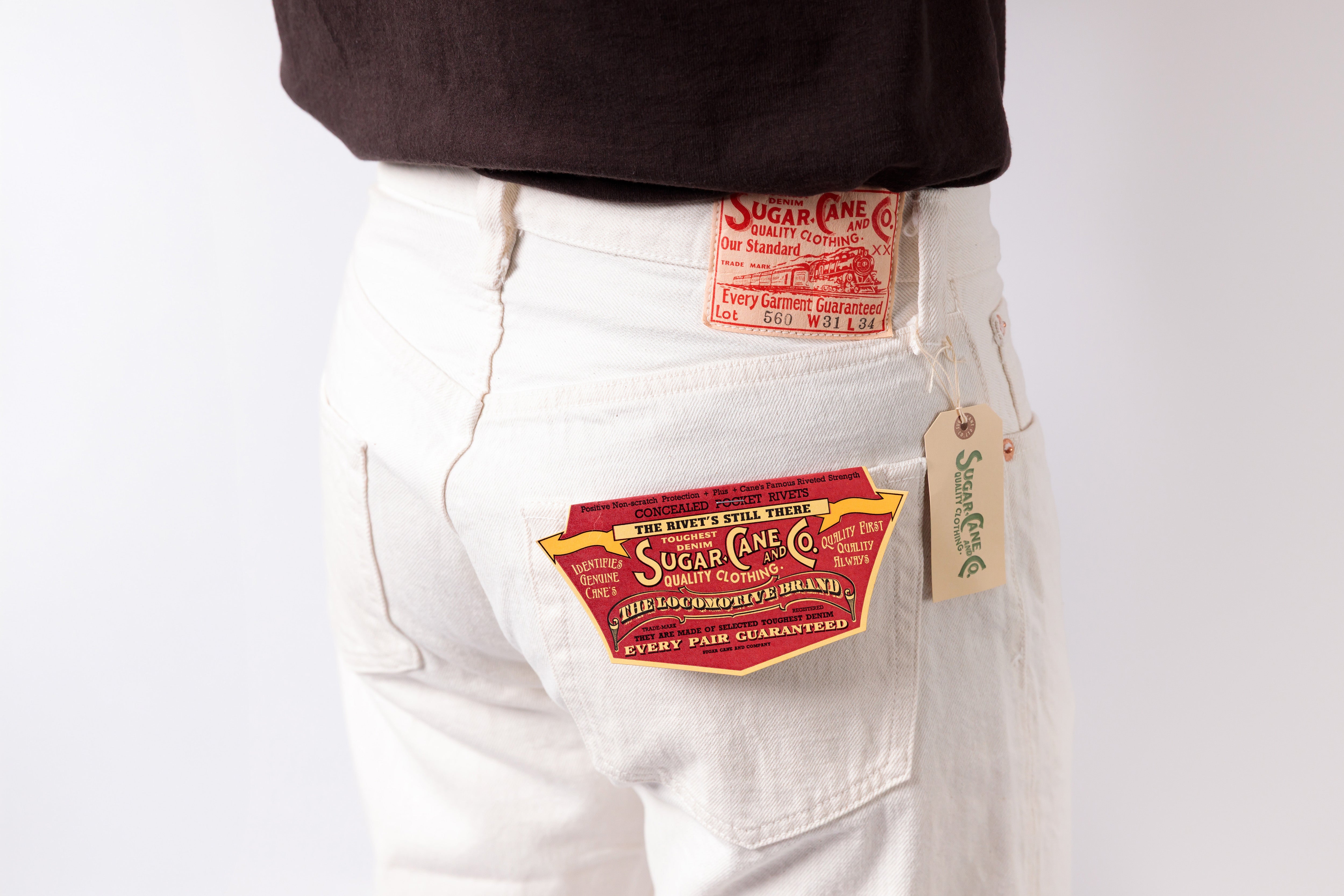 Close-up of a person wearing white denim jeans with multiple Sugar Cave brand patches on the back pocket and waistband, paired with a dark brown top against a plain white background.