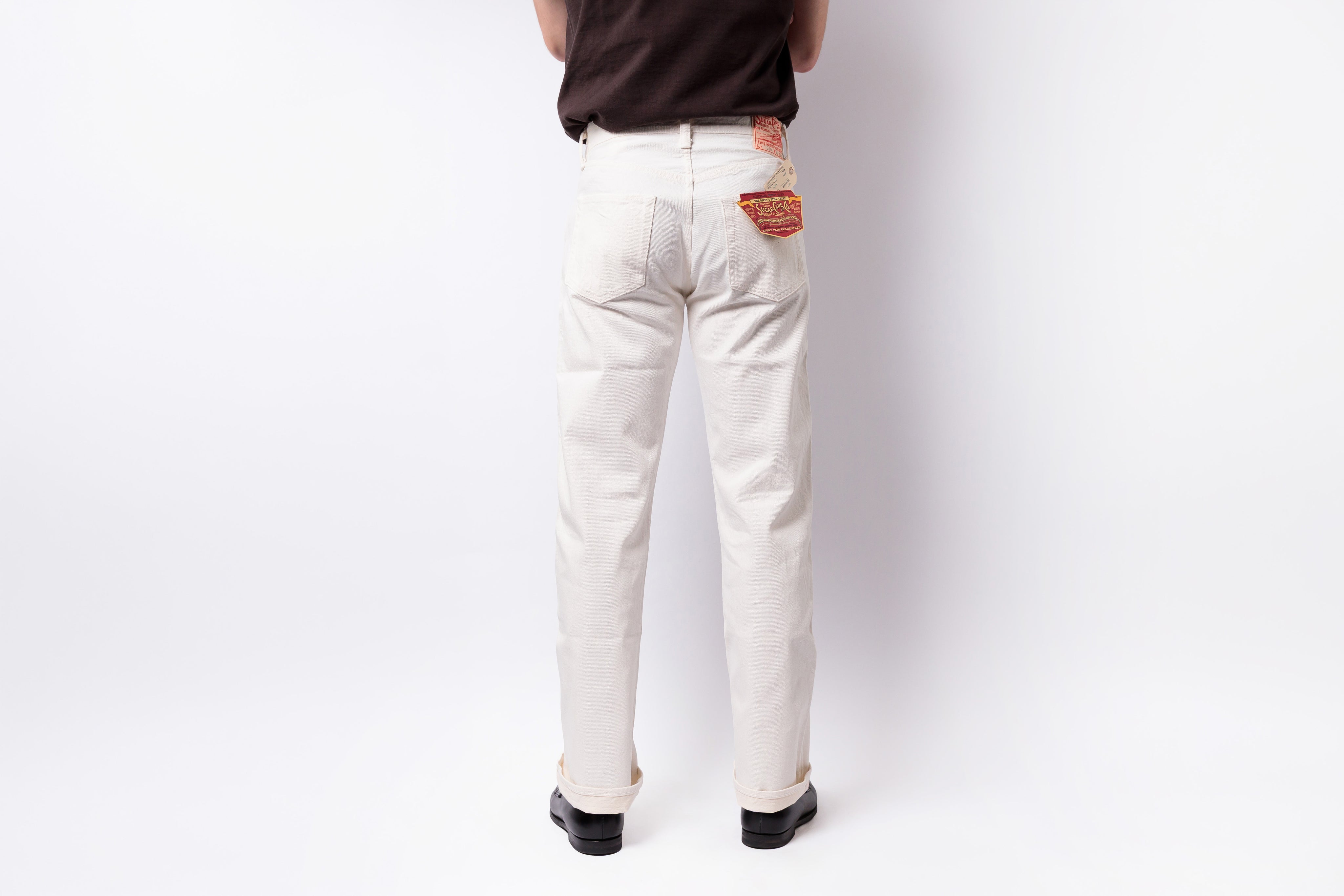 A person shown from the back wearing off-white denim jeans with a brown shirt and black shoes, standing against a plain white studio background.