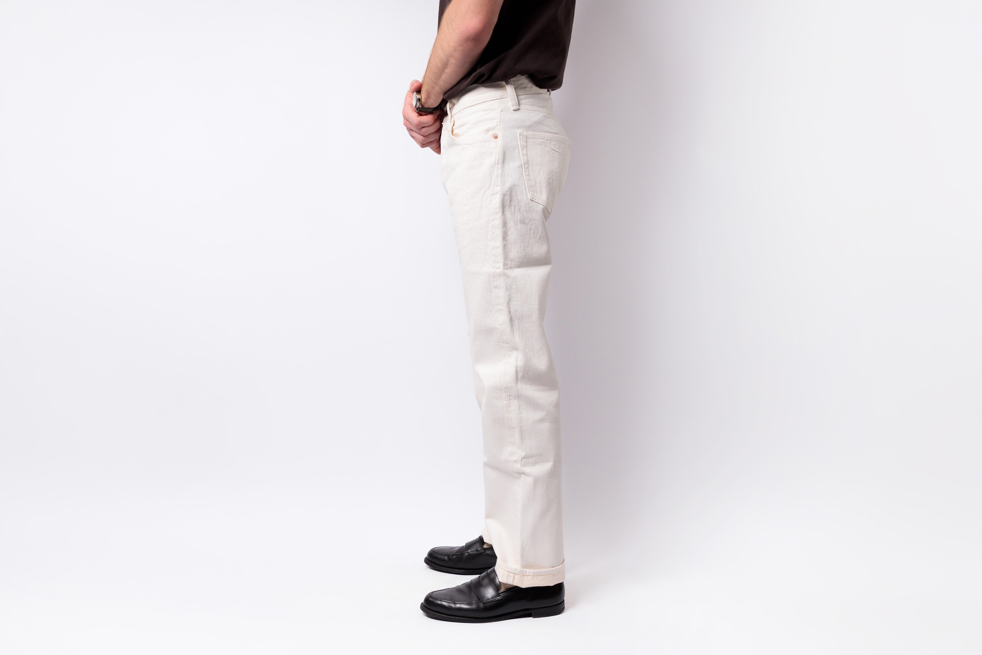 A side profile of a person wearing off-white straight-leg denim pants, a dark brown t-shirt, and black leather loafers, standing against a plain white studio background.