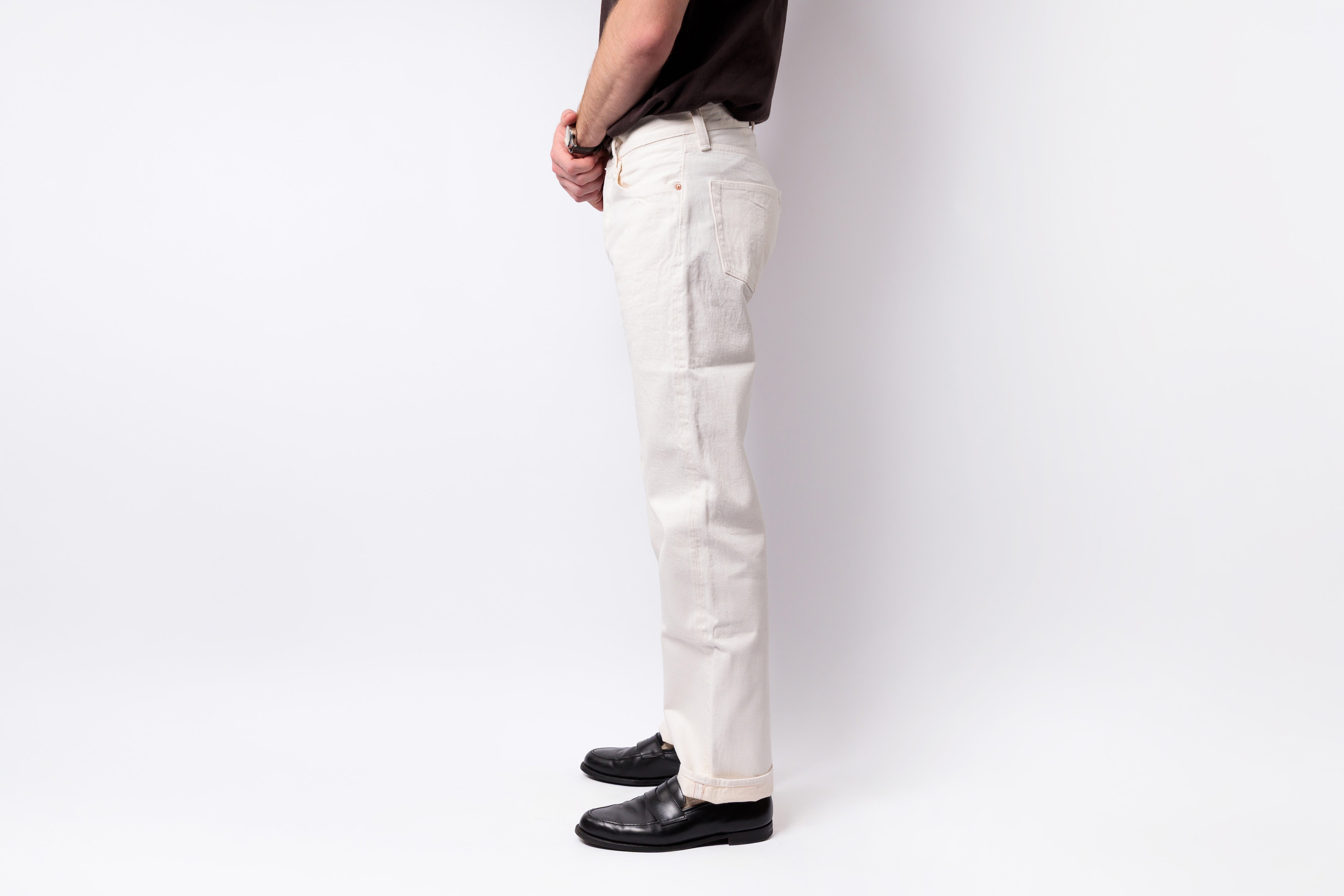 A side profile of a person wearing off-white straight-leg denim pants, a dark brown t-shirt, and black leather loafers, standing against a plain white studio background.