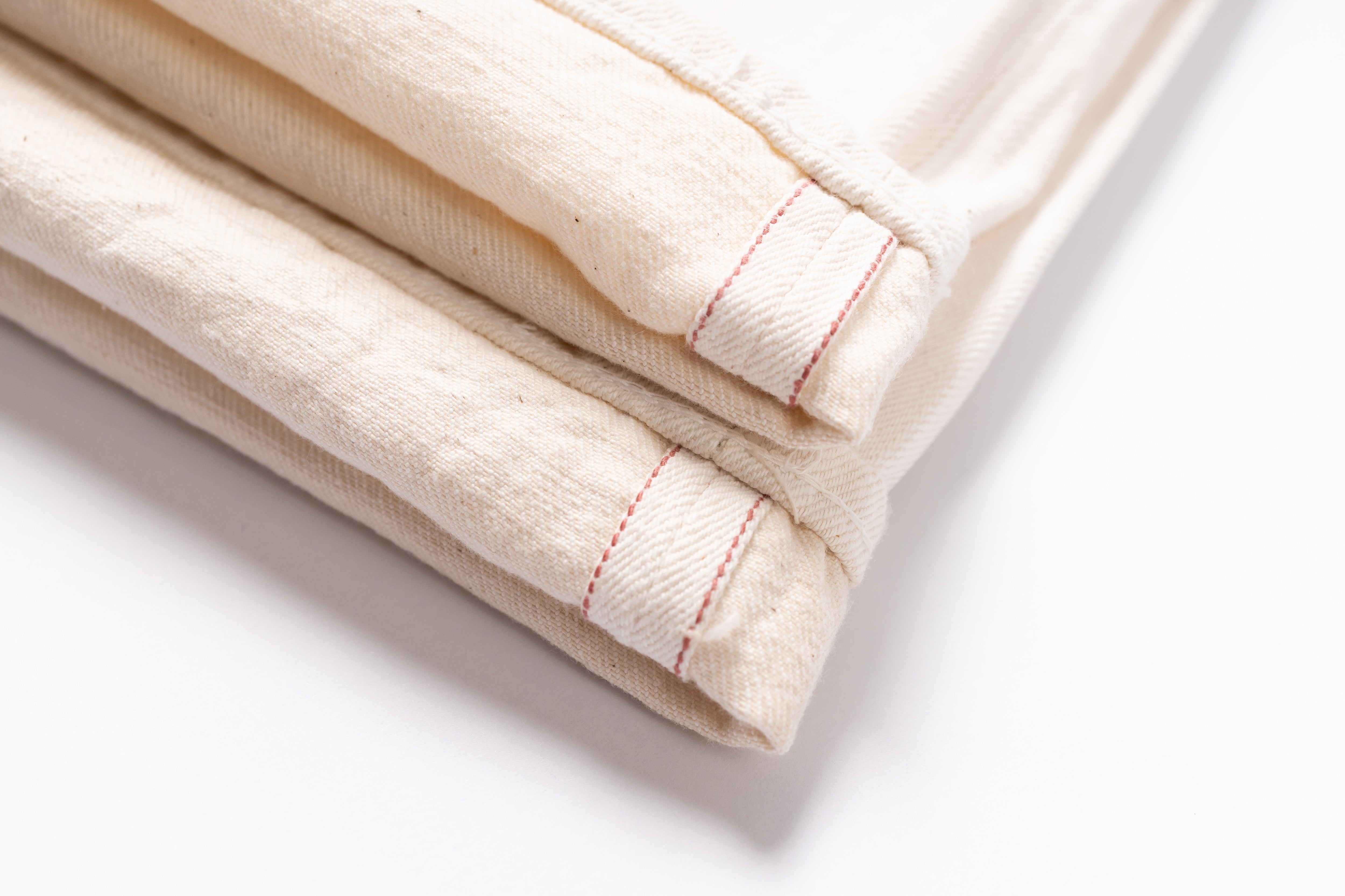 A close-up of folded off-white fabric with visible red stitching along the edges, photographed on a clean white background.