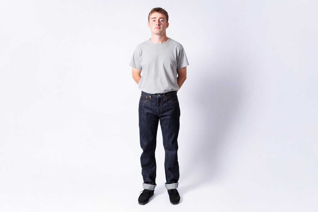 Man wearing a gray t-shirt and dark jeans standing against a white background