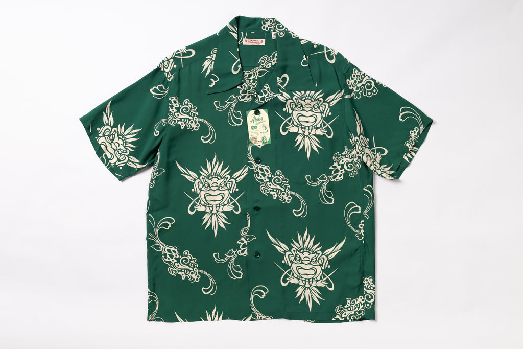 A short-sleeve button-up shirt in dark green with a repeating cream-colored graphic print of stylized faces and ornamental swirls, laid flat against a white background. A hangtag is attached near the collar.