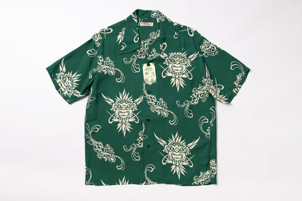 A short-sleeve button-up shirt in dark green with a repeating cream-colored graphic print of stylized faces and ornamental swirls, laid flat against a white background. A hangtag is attached near the collar.