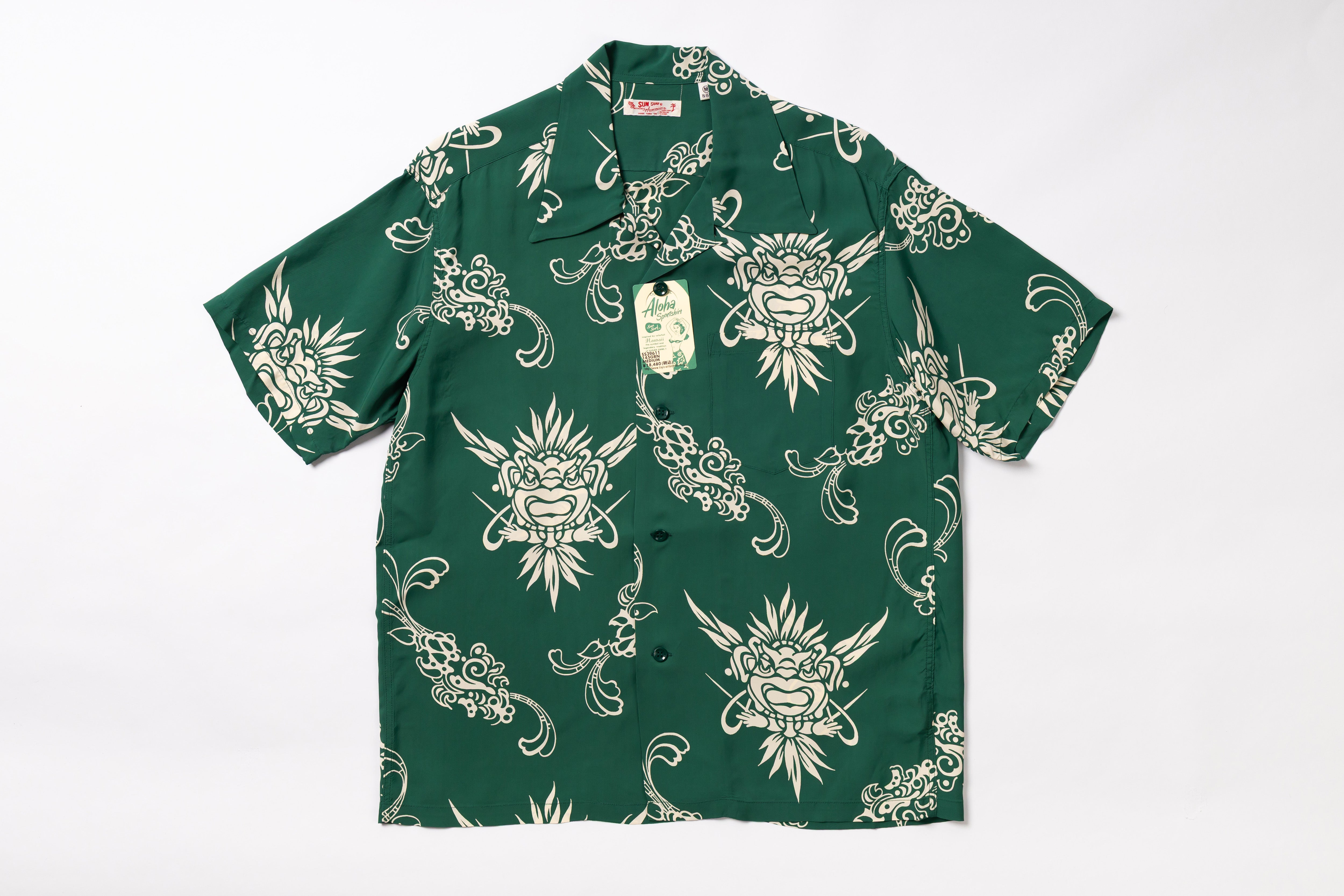 A short-sleeve button-up shirt in dark green with a repeating cream-colored graphic print of stylized faces and ornamental swirls, laid flat against a white background. A hangtag is attached near the collar.