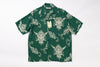 A short-sleeve button-up shirt in dark green with a repeating cream-colored graphic print of stylized faces and ornamental swirls, laid flat against a white background. A hangtag is attached near the collar.