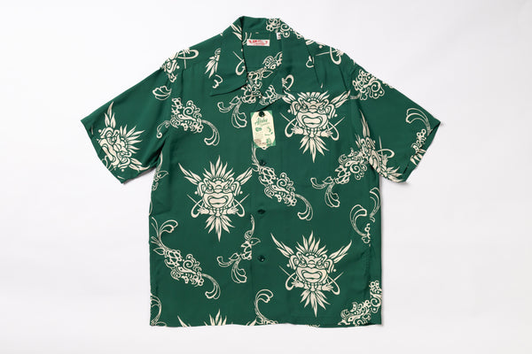 A short-sleeve button-up shirt in dark green with a repeating cream-colored graphic print of stylized faces and ornamental swirls, laid flat against a white background. A hangtag is attached near the collar.