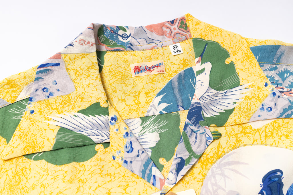 A close-up of a yellow short-sleeve camp collar shirt with a Japanese-style crane print in green, blue, and white, featuring a visible 'Sun Surf' brand label.