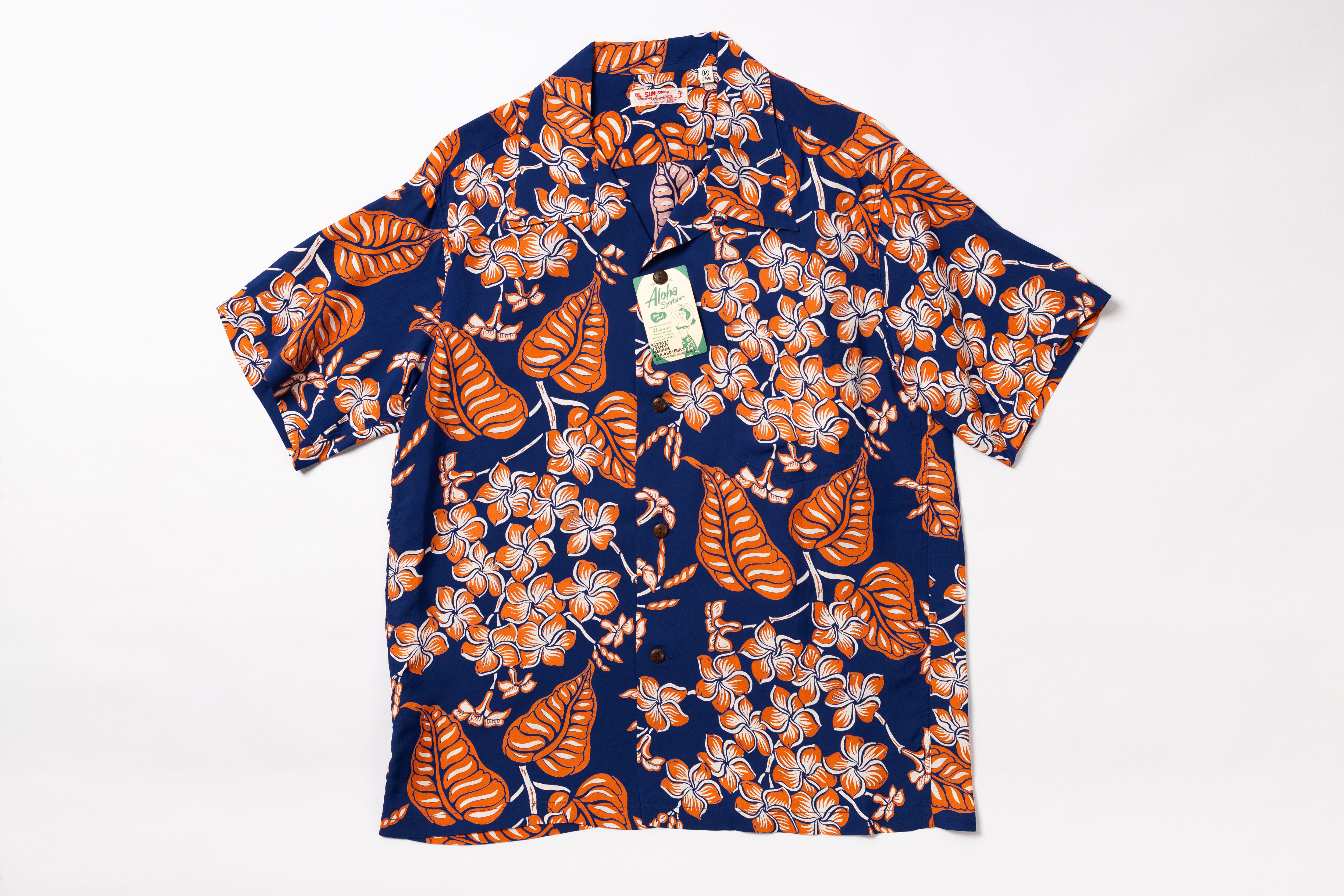 A short-sleeve button-up camp shirt with a navy blue base, featuring a bold orange tropical leaf and white plumeria floral pattern, laid flat on a white background with a paper tag attached.