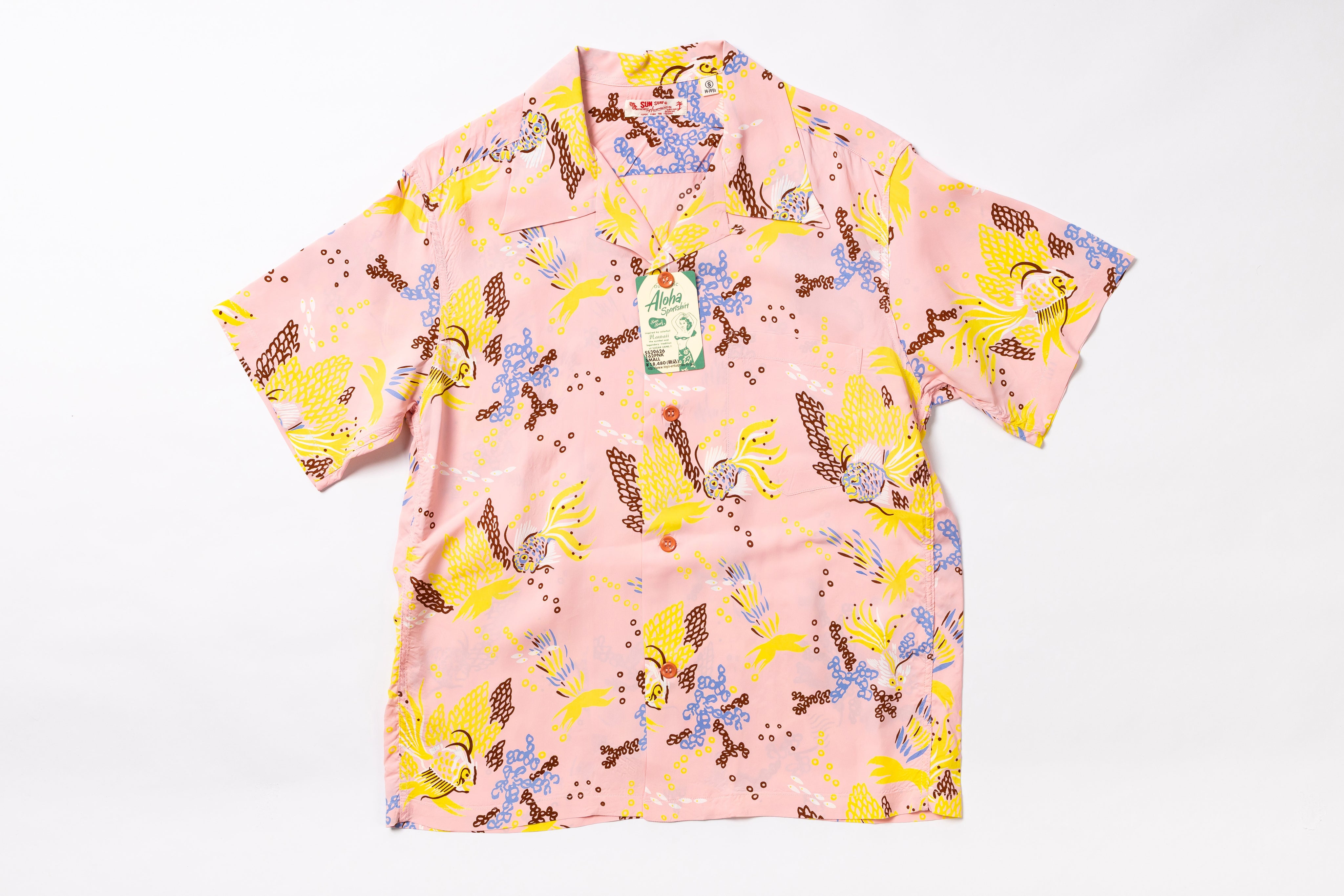 A short-sleeve collared button-up shirt in light pink with a colorful cartoon-style all-over print featuring yellow, blue, brown, and purple elements. A hangtag is attached at the collar, and the shirt is photographed flat against a plain white background.