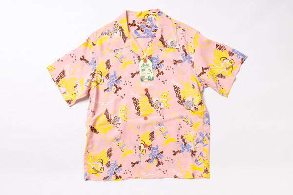 A short-sleeve collared button-up shirt in light pink with a colorful cartoon-style all-over print featuring yellow, blue, brown, and purple elements. A hangtag is attached at the collar, and the shirt is photographed flat against a plain white background.