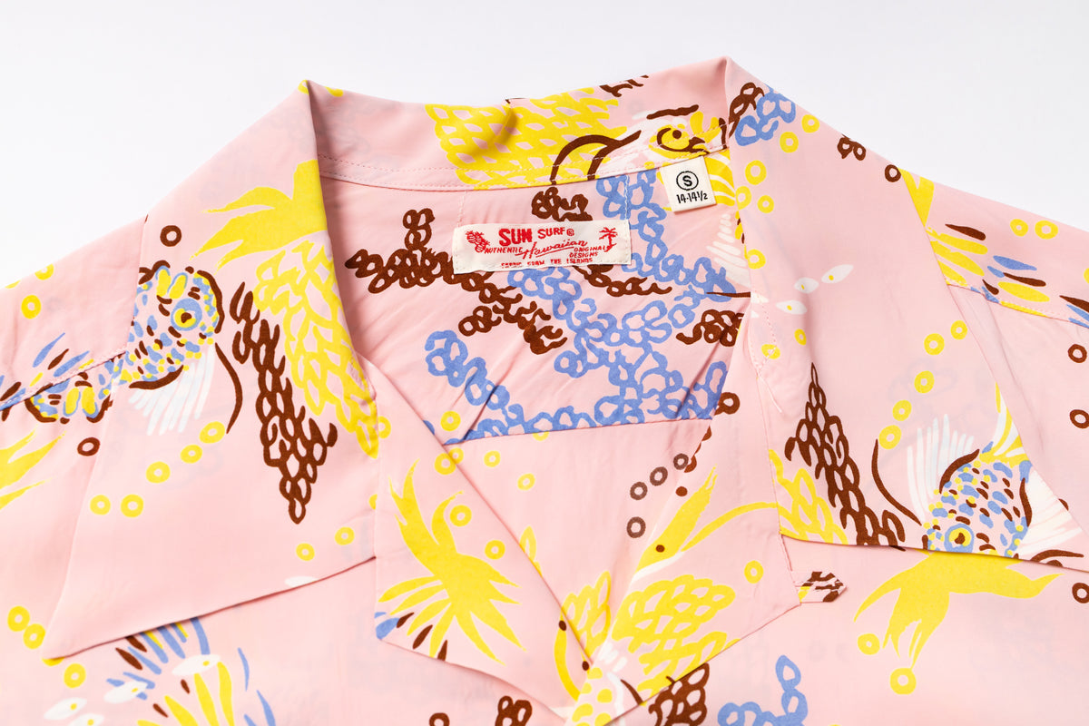 A close-up of a pink short-sleeve camp collar shirt with a colorful print of yellow, blue, and brown tropical birds and abstract patterns. The brand label 'Sun Surf' is visible inside the collar.