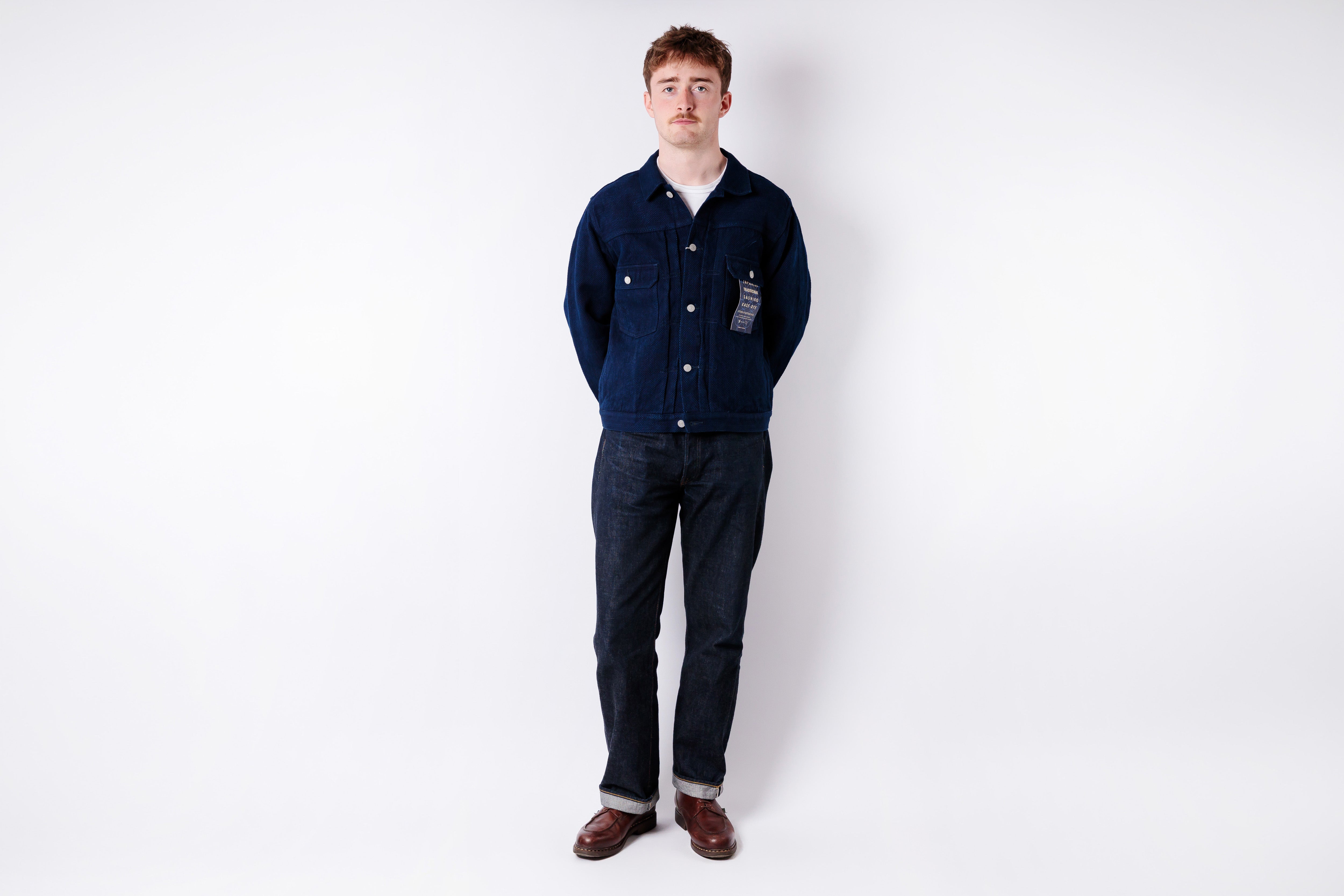 Man wearing a navy blue jacket and dark jeans standing against a white background