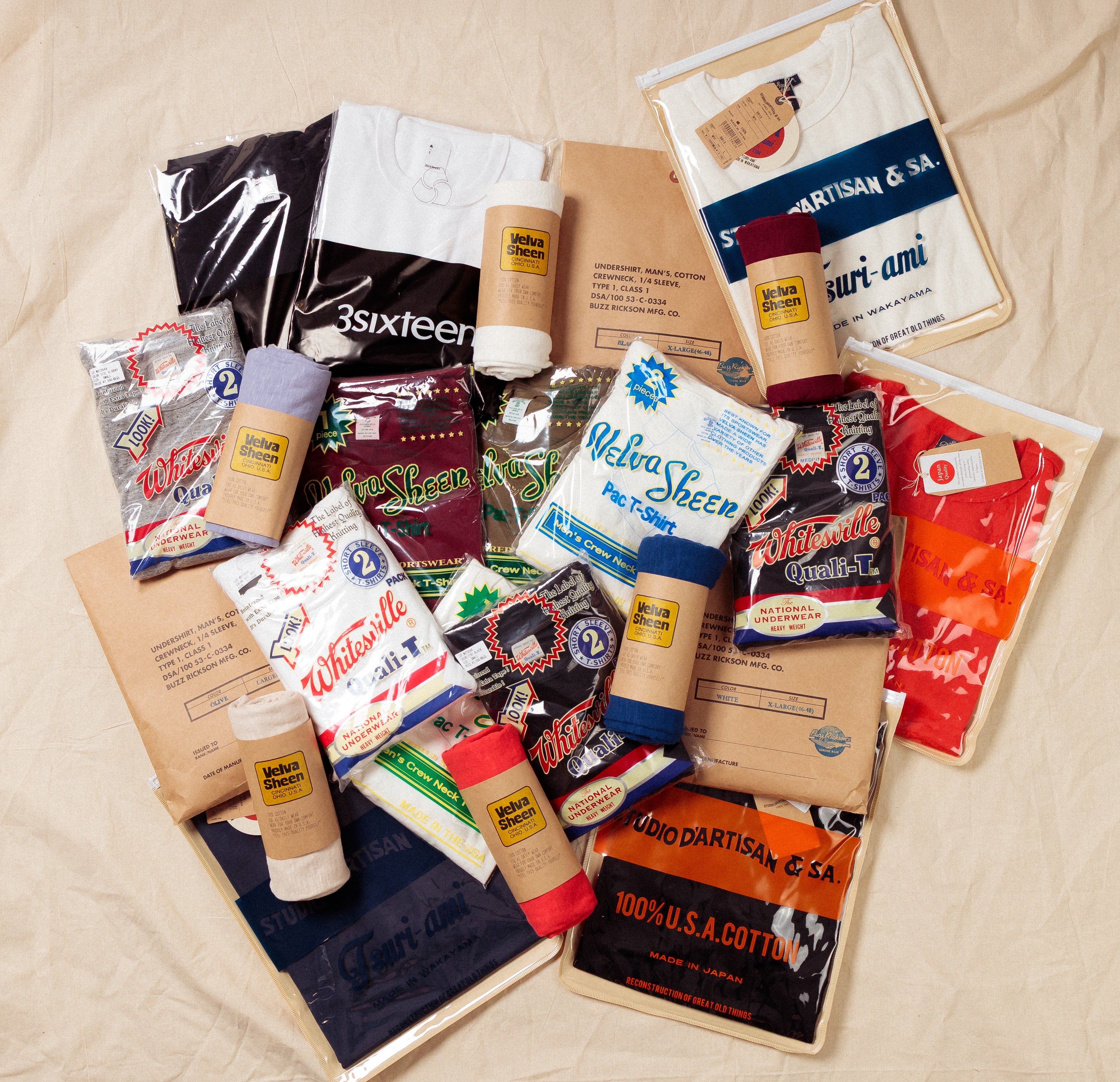 A curated gift bundle of cotton apparel and snacks arranged on a beige fabric background. The bundle includes multiple t-shirts in various colors, several brown kraft paper gift boxes, and assorted packaged snacks such as chips, pretzels, and candy.