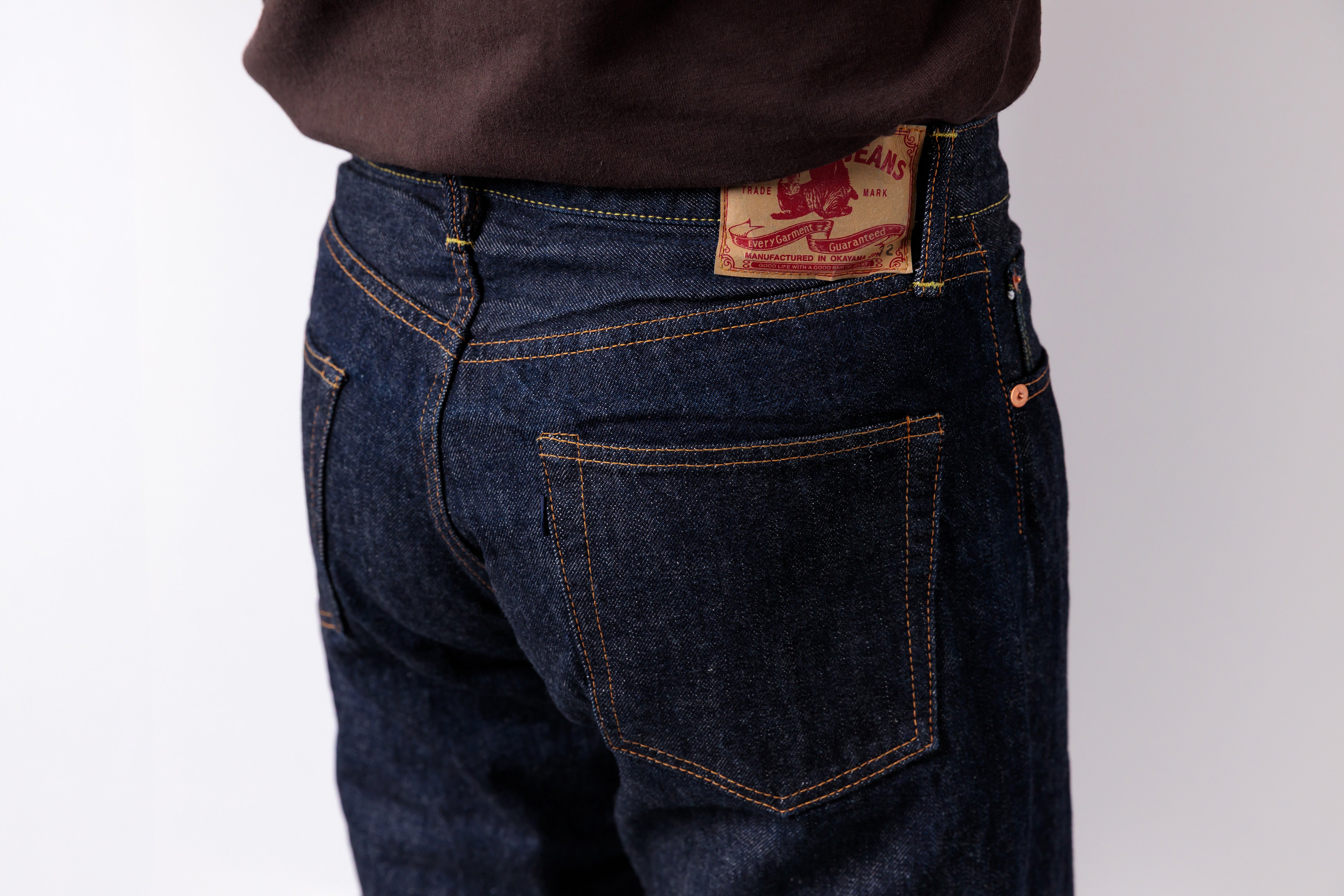 Close-up back view of a person wearing dark indigo jeans with a brown top, showing the back pocket and waistband patch.