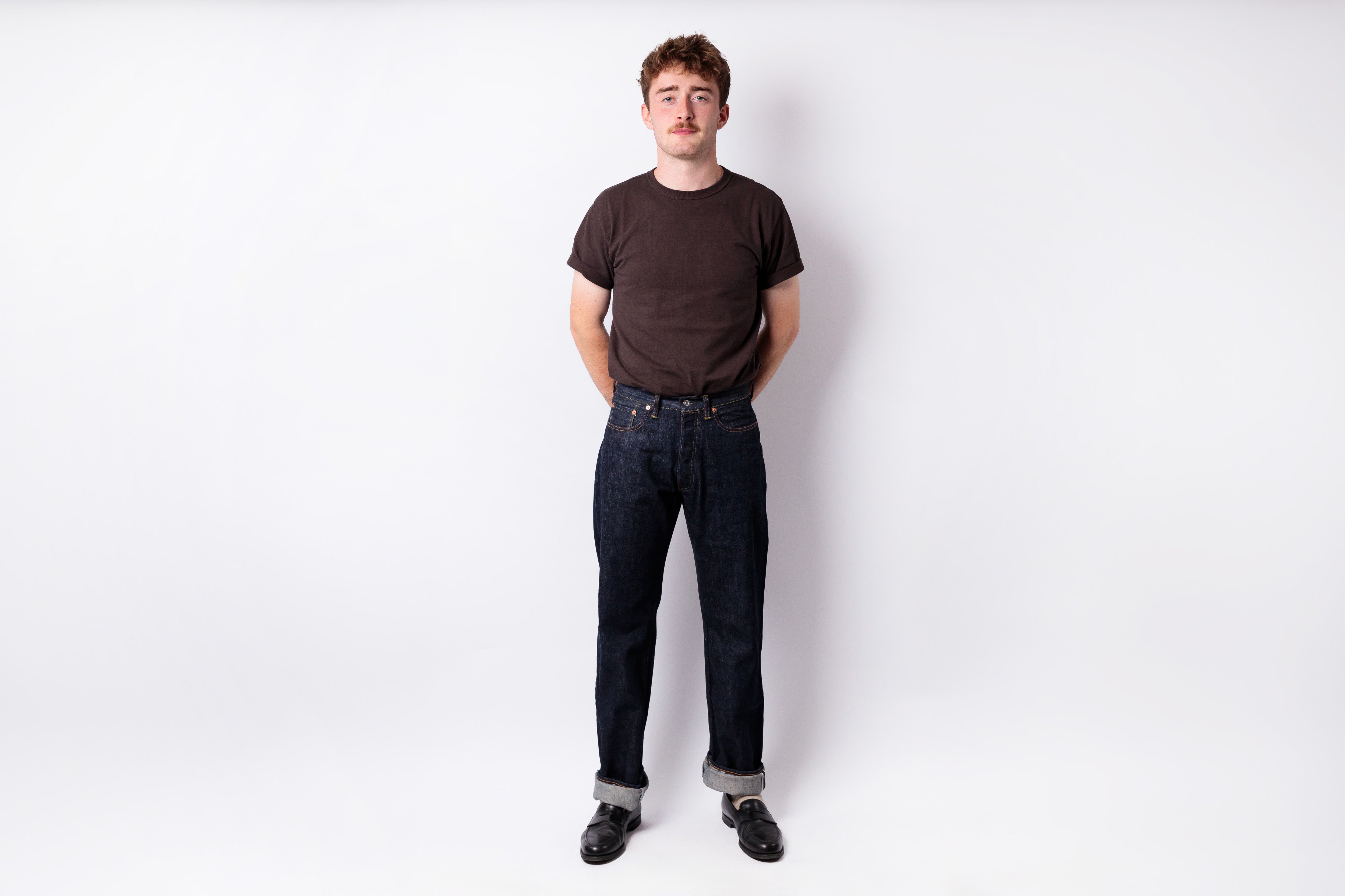 A man standing against a white studio background wearing a dark brown crewneck t-shirt, dark blue jeans, and black shoes.
