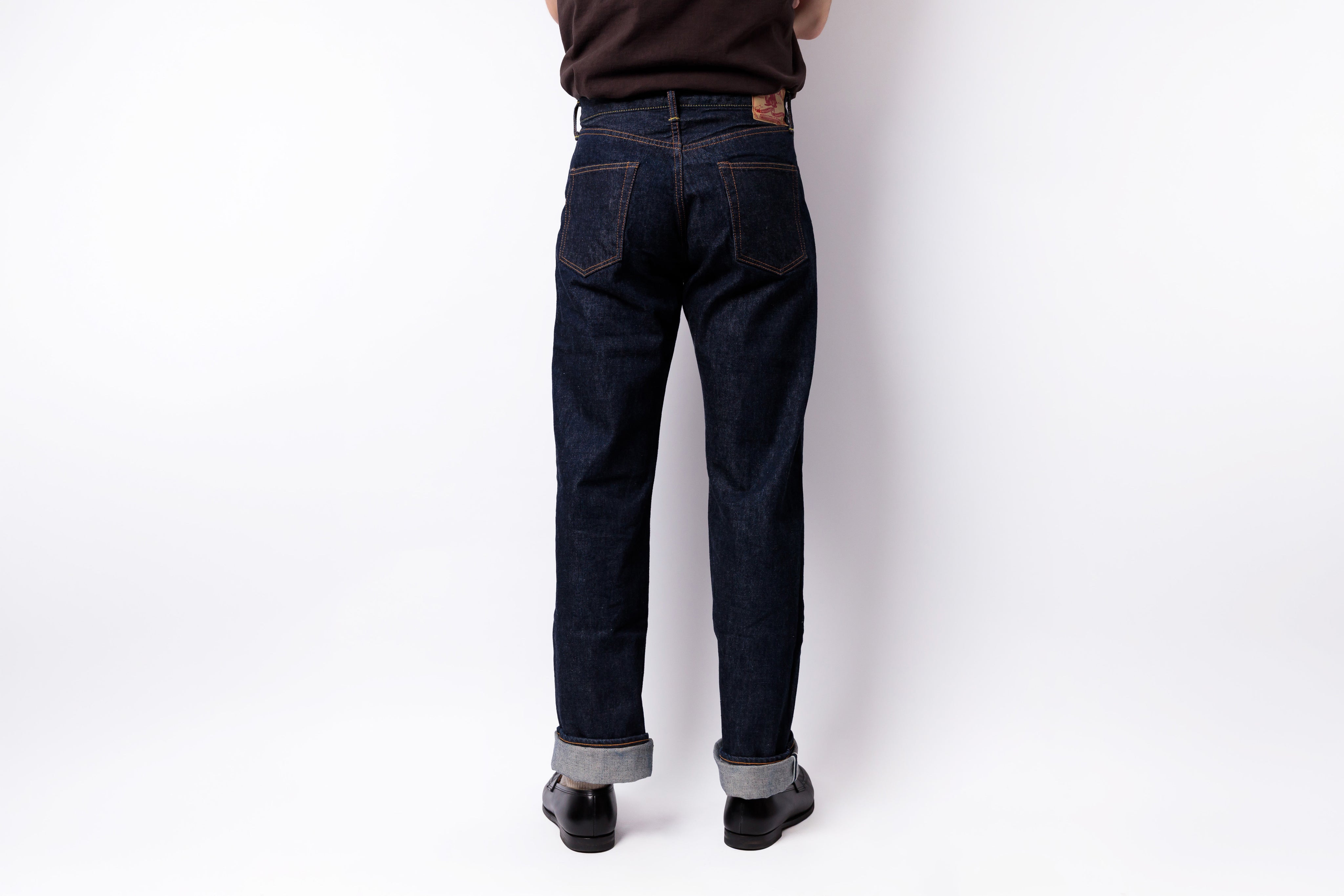 A person shown from the back wearing dark indigo denim jeans with rolled cuffs, black shoes, and a dark brown top, standing against a plain white studio background.