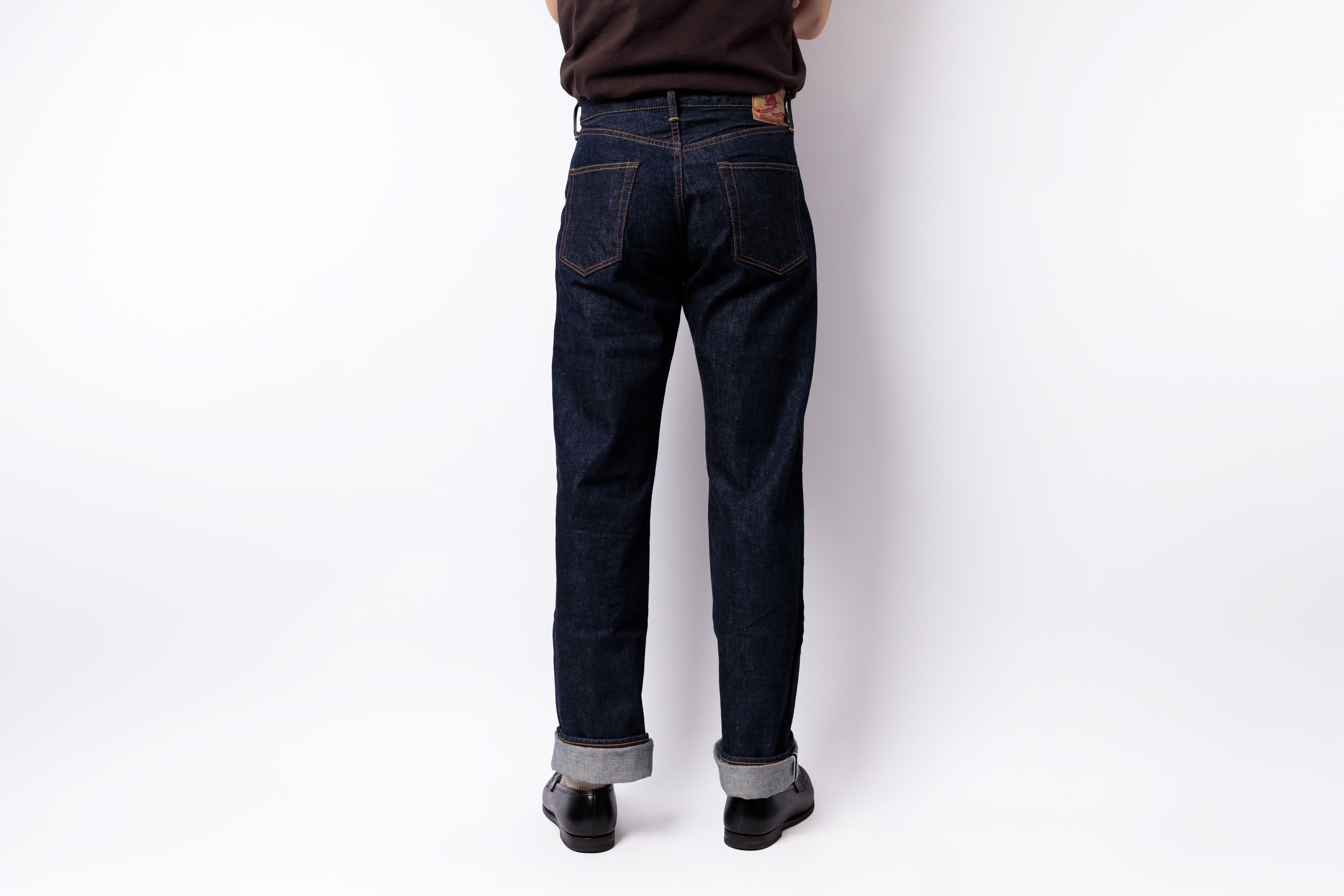 A person shown from the back wearing dark indigo denim jeans with rolled cuffs, black shoes, and a dark brown top, standing against a plain white studio background.